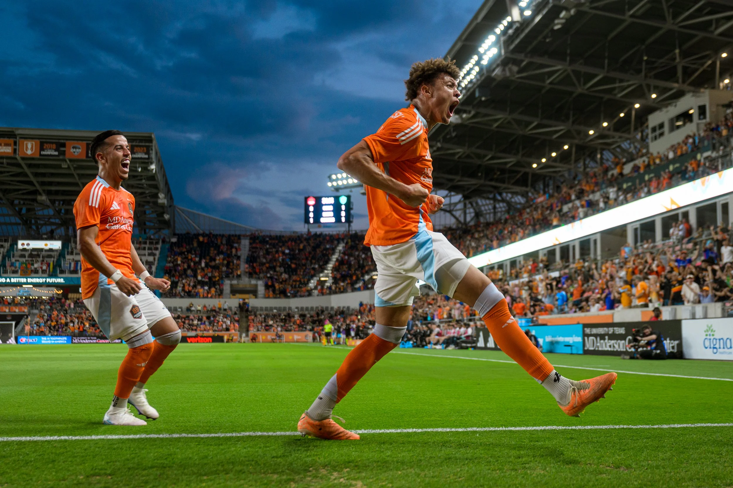 Two soccer players in orange jerseys celebrating on the field at a stadium, with a large crowd in the stands and a digital scoreboard in the background.