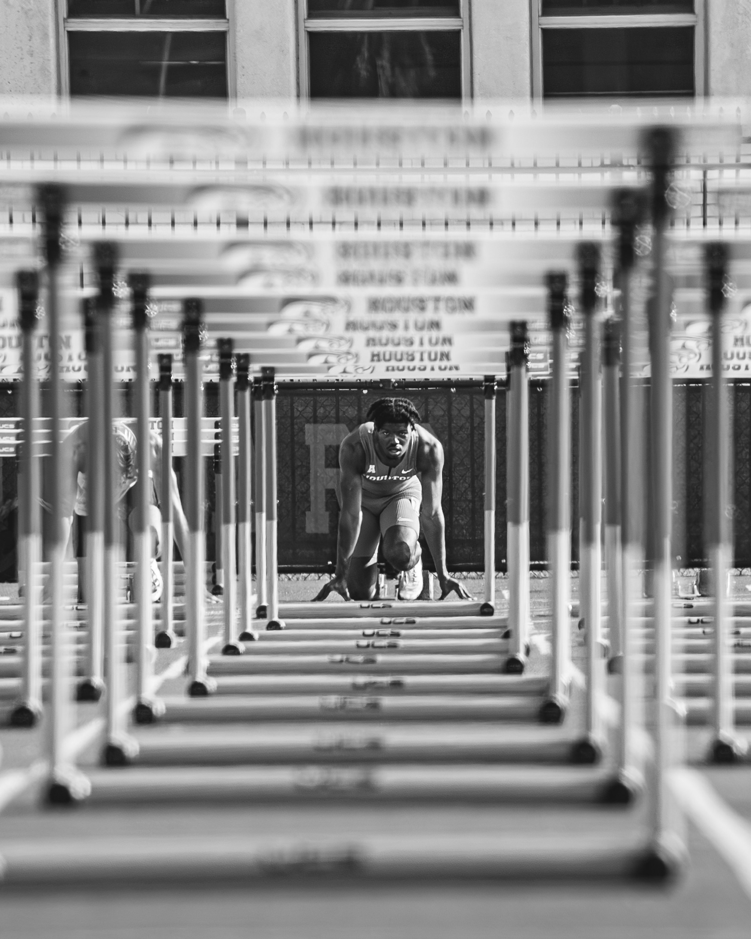 A male athlete in a starting position behind hurdles on a track, preparing to race, with a fence and building in the background.