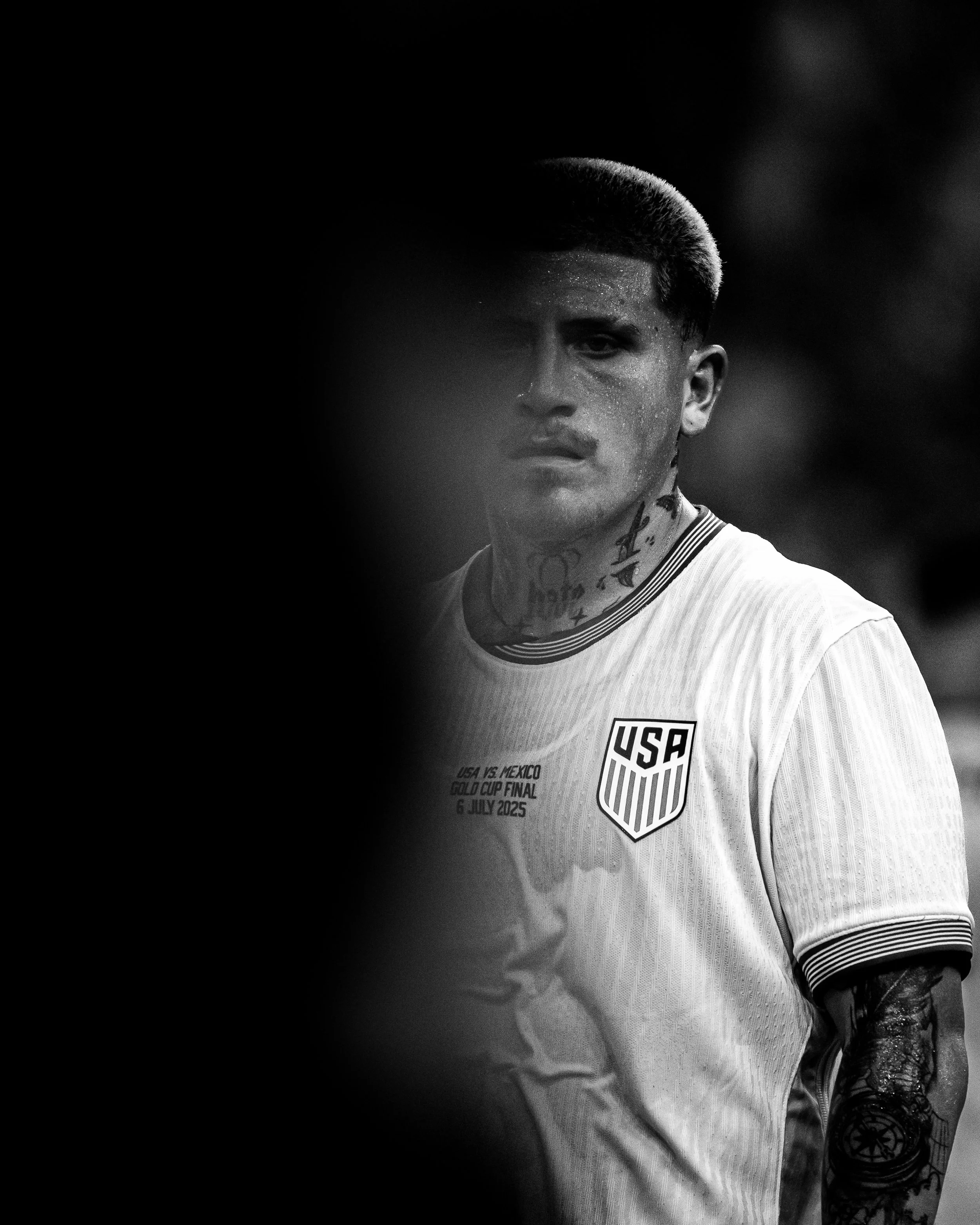 A black and white photo of a male soccer player with tattoos, wearing a USA national team jersey with the text "USA vs. Mexico Gold Cup Final 6 July 2025." The player has short hair and a serious expression, with part of the image obscured on the lef