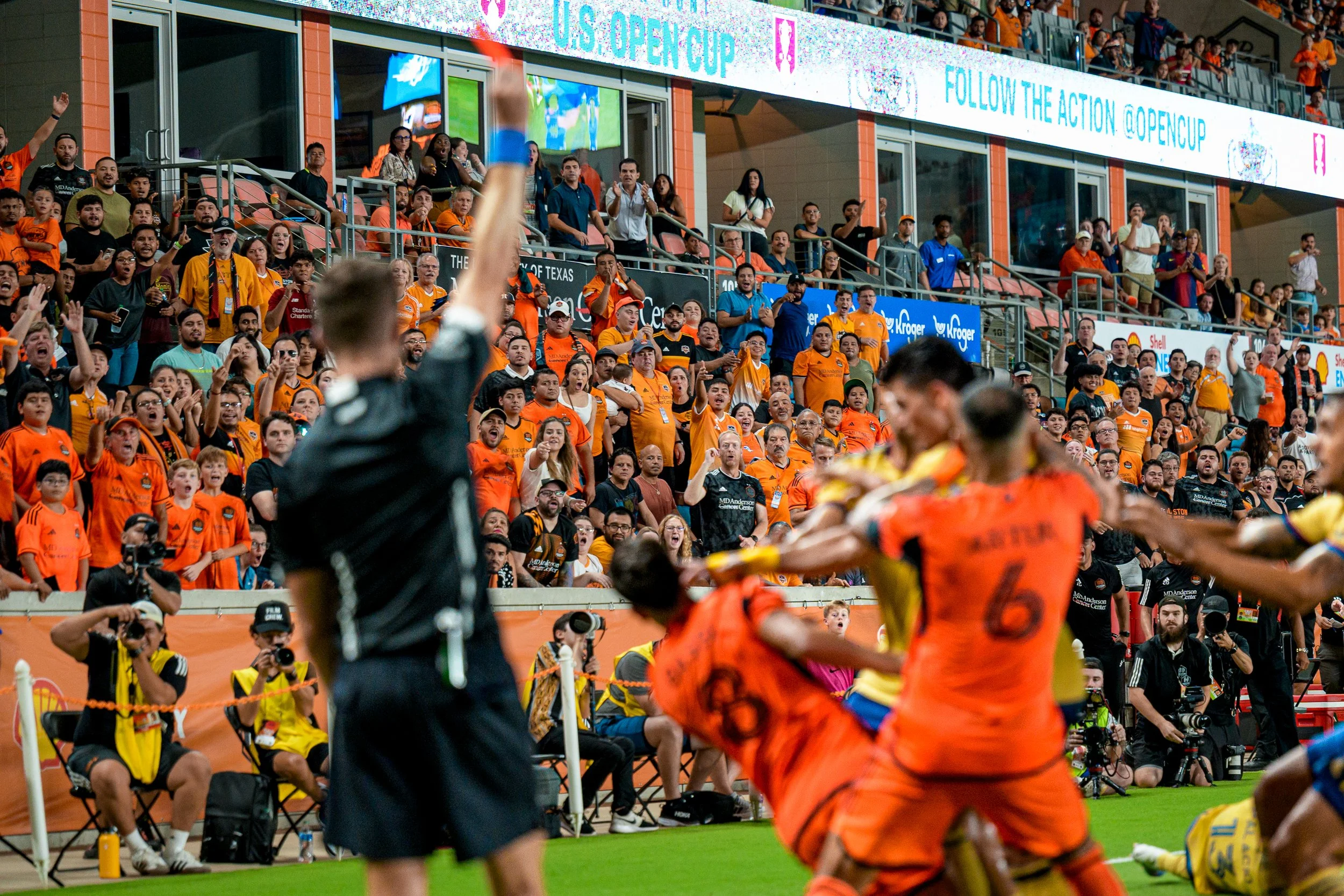 Soccer players in orange and yellow uniforms on the field as a referee raises his arm, with a crowd of fans dressed in orange cheering in the background at a stadium.