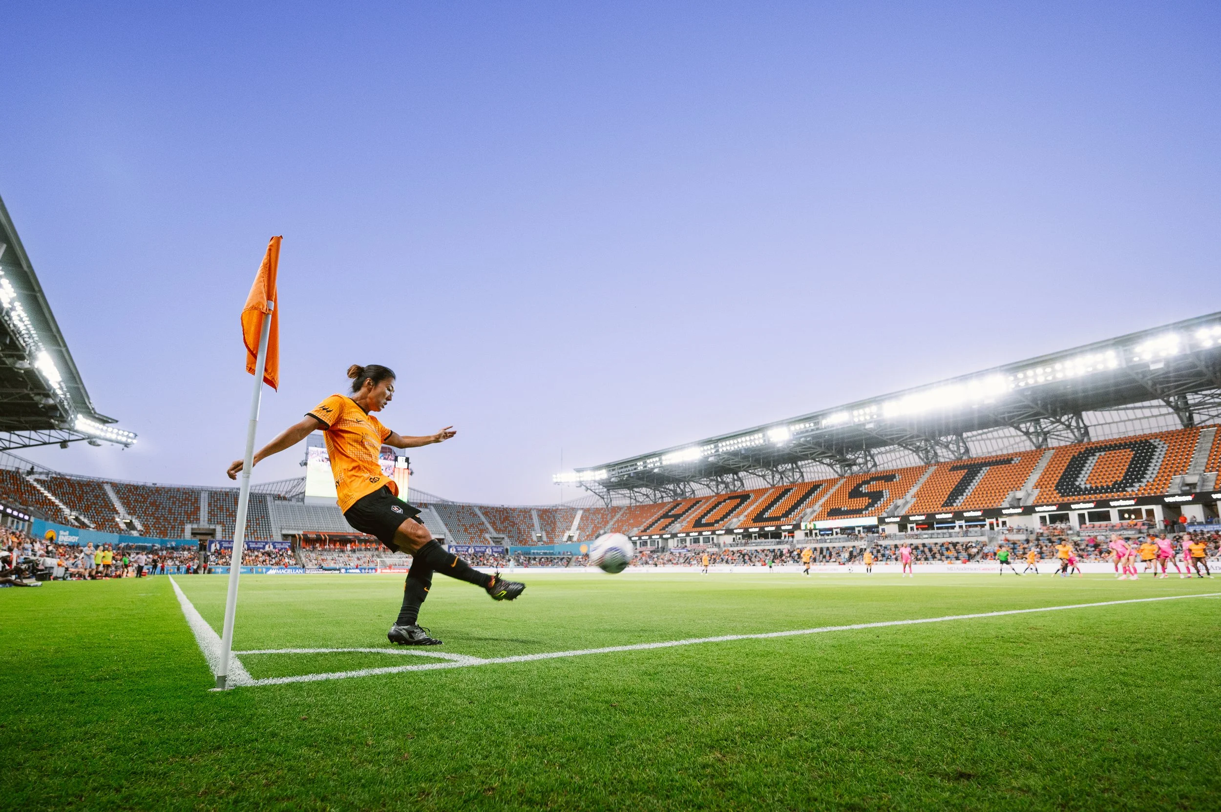 Female soccer player taking a corner kick on a soccer field during a game at a large stadium with the word 'HOUSTON' on the stands.
