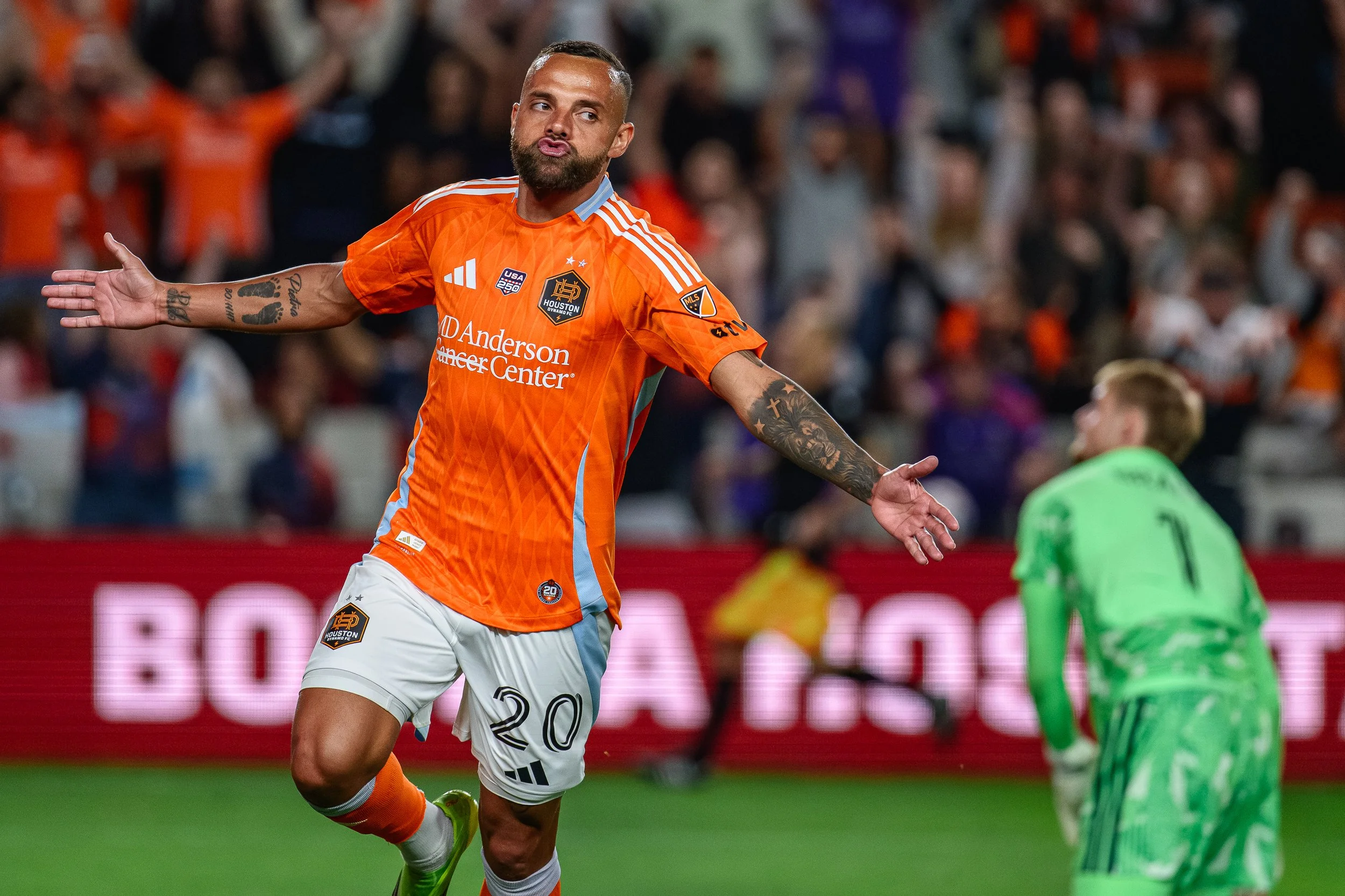A soccer player celebrating with arms open during a match. He is wearing an orange jersey with the number 20, white shorts, and bright yellow shoes. A goalkeeper in a green uniform is in the background.