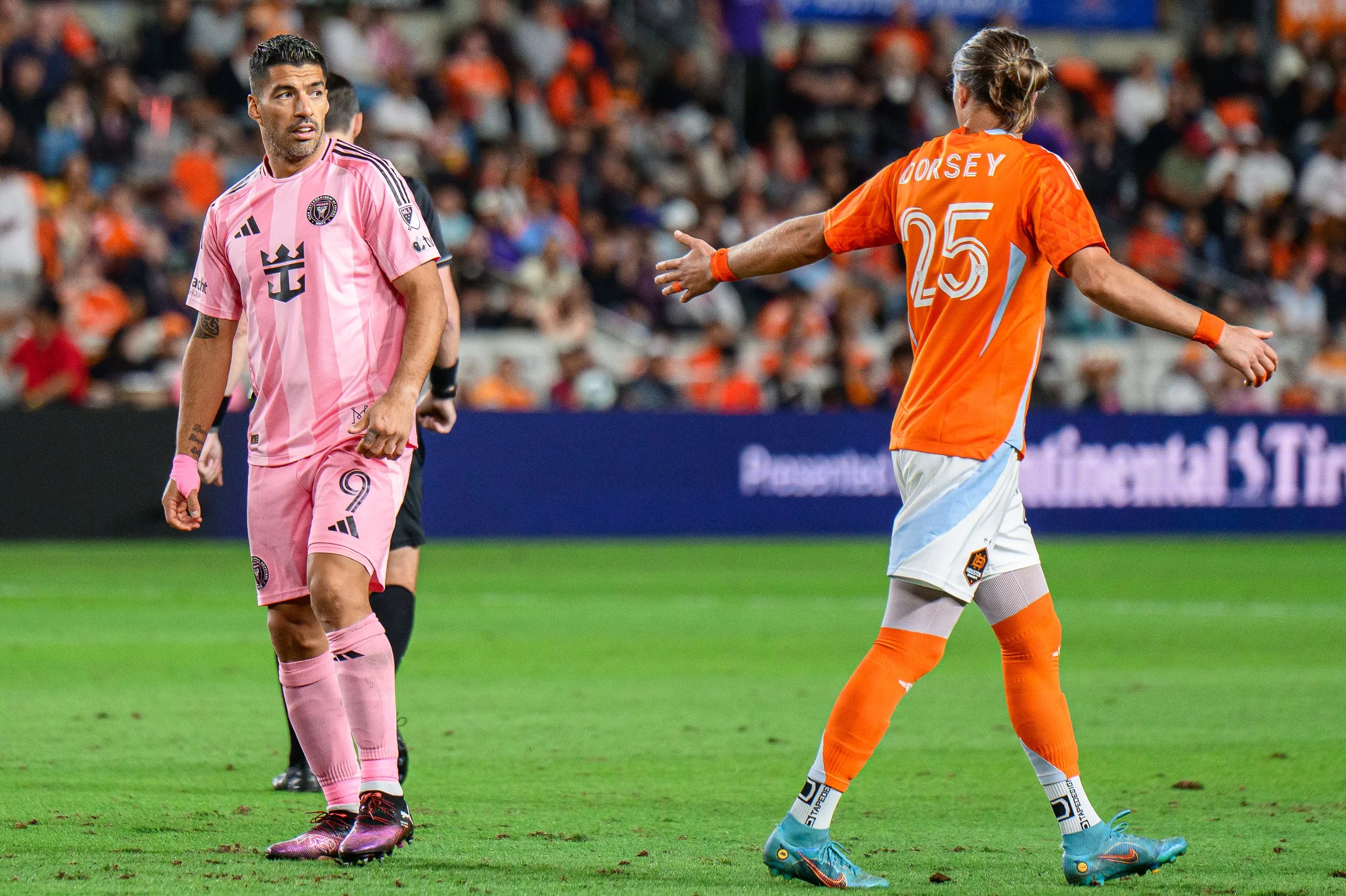 Two soccer players on the field during a match, one in pink and the other in orange, with a crowd in the background.