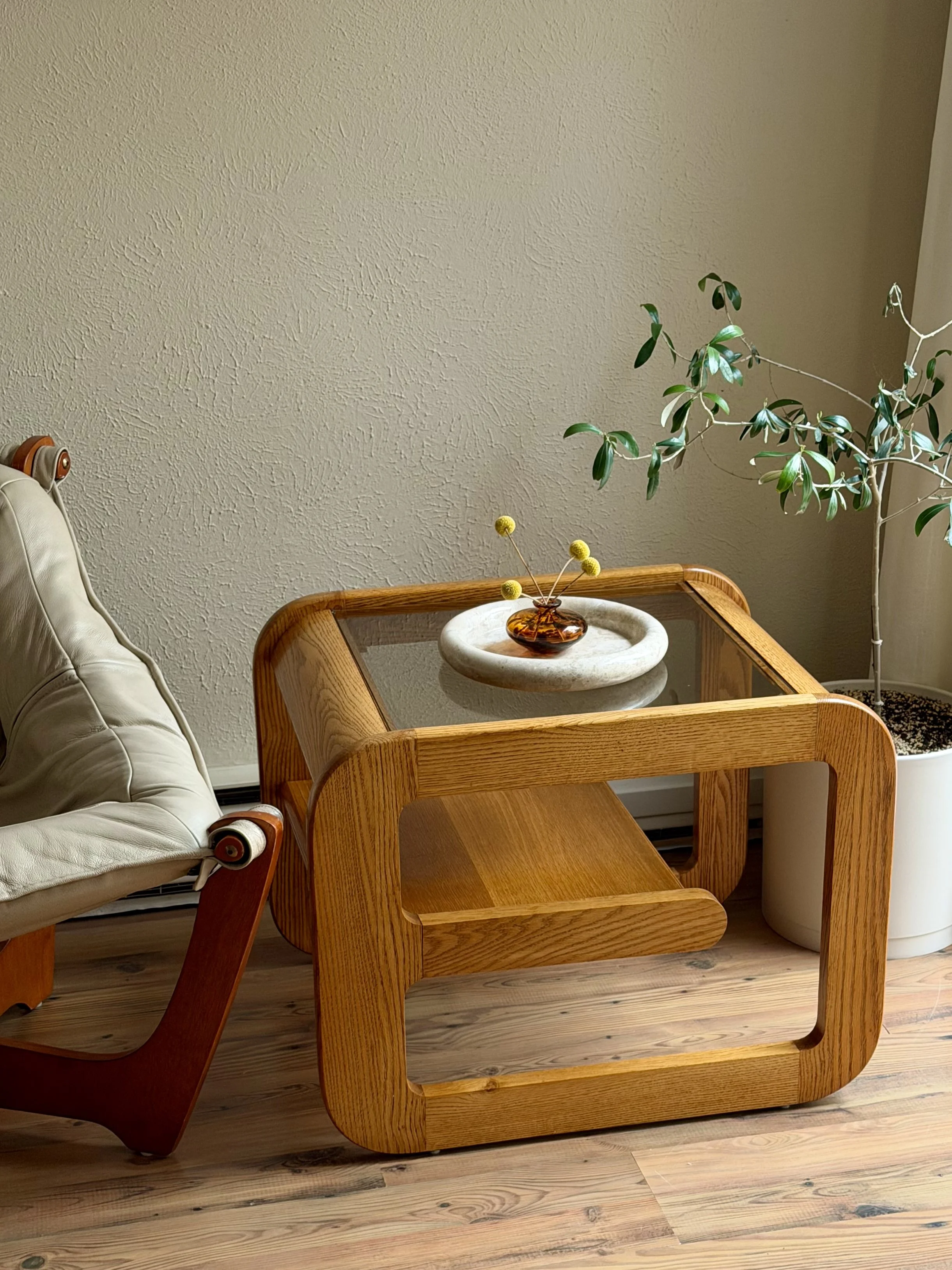 A wooden side table with a round, white tray holding a small amber glass vase with yellow flower stems, next to a large potted plant with green leaves, in a room with wooden flooring and textured beige walls.