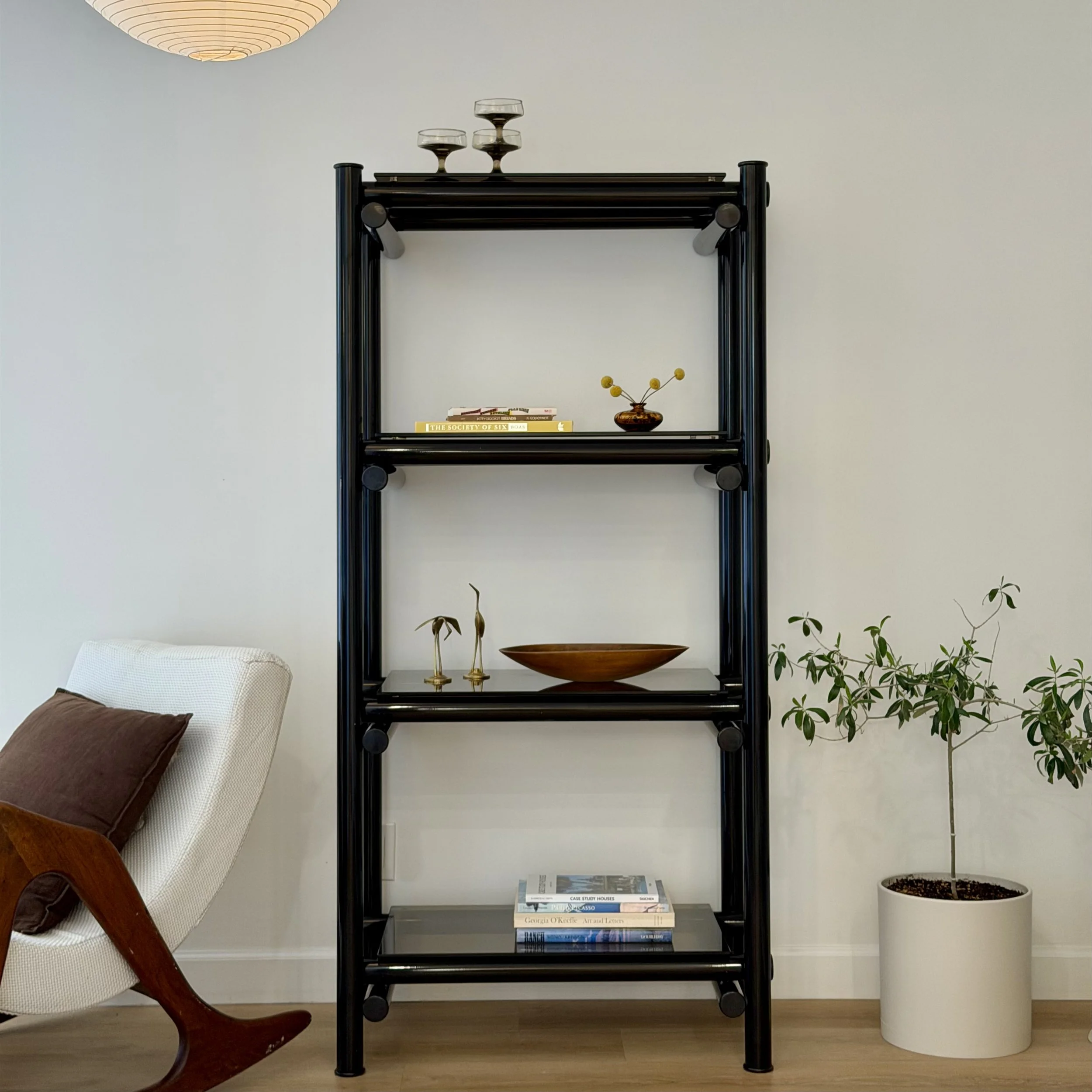 Black metal bookshelf with three shelves containing books, a small vase with yellow flowers, and a decorative bowl. To the left, part of a white armchair with a brown cushion and wooden armrest. To the right, a tall potted plant. A paper or magazine 