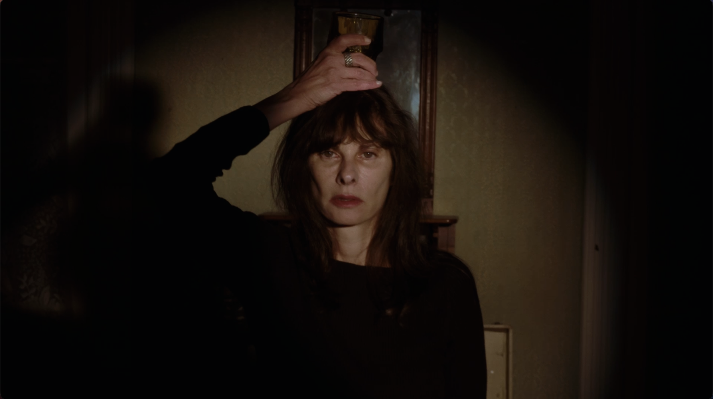 A woman with brown hair and a black top is holding a glass of drink on her head in a dimly lit room.