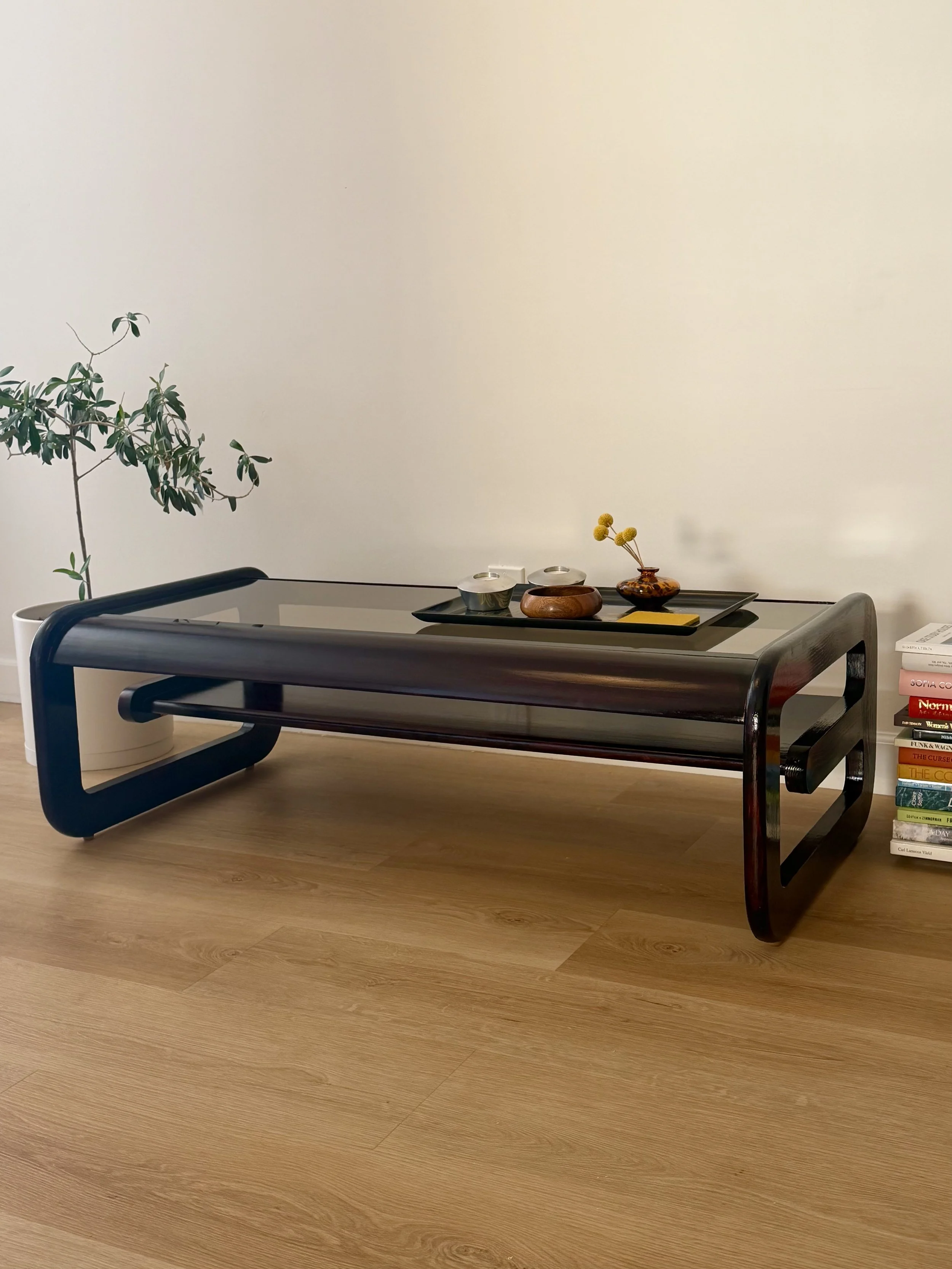 A modern coffee table with a black frame and wooden surface, placed on a light wooden floor, with a black tray holding small decorative vases and yellow flowers, a stack of books on the side, and a white potted plant in the background against a plain
