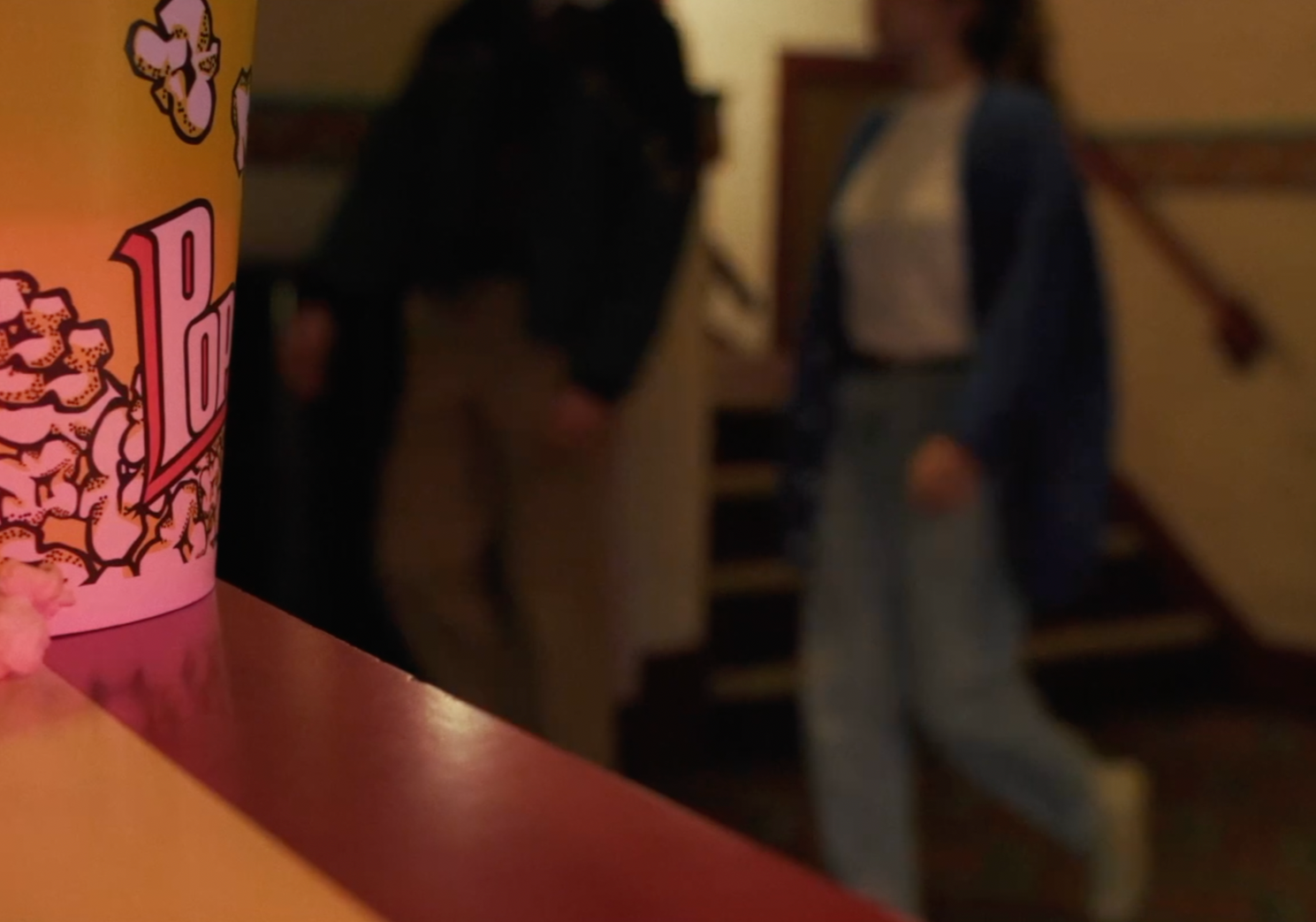 Close-up of a pink popcorn bucket with the word "Pop" on it, in focus. In the blurred background, two women are seen standing on a staircase in an indoor setting.