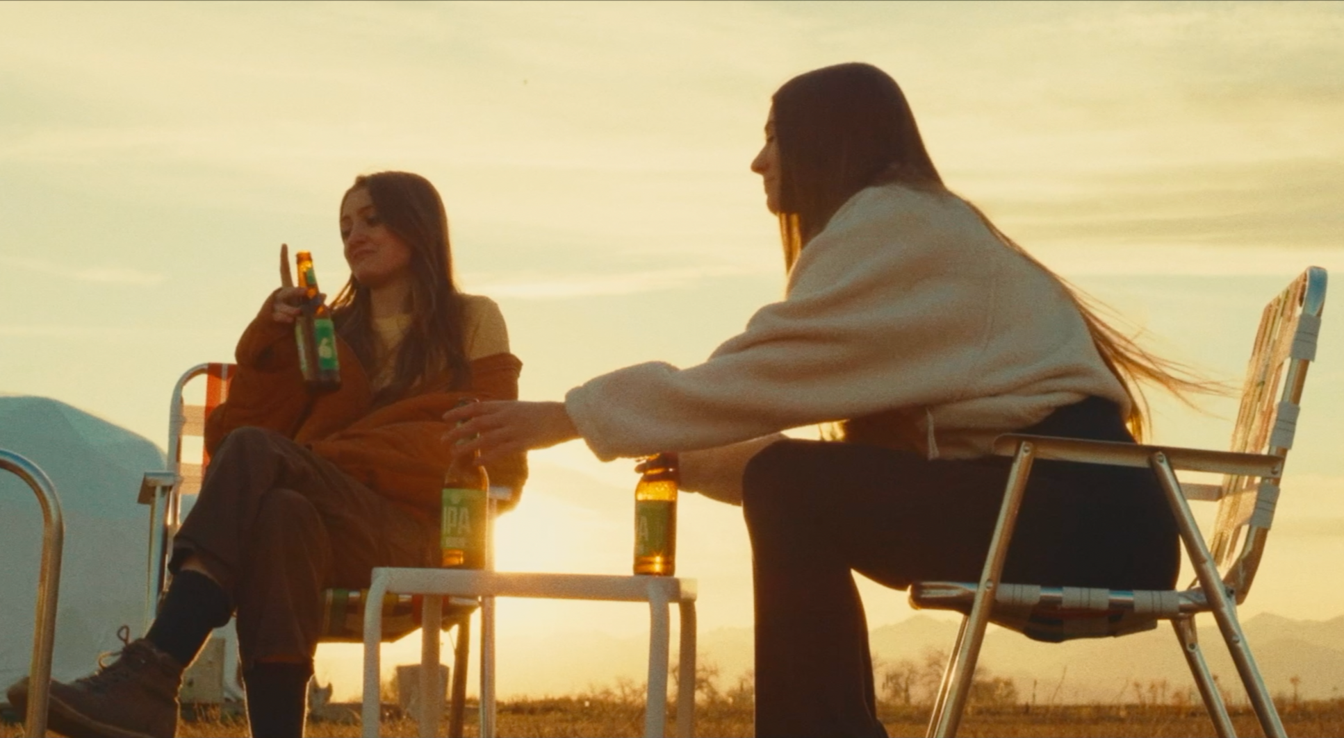 Two young women sitting outdoors during sunset, drinking bottled beverages, sitting on camping chairs with relaxed expressions.