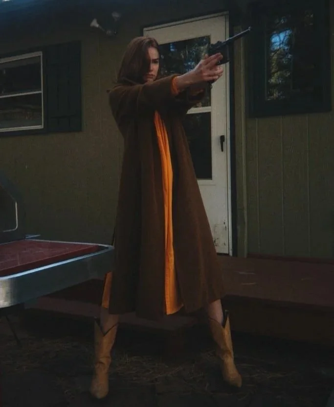 A woman standing outside, aiming a handgun, wearing cowboy boots and a long brown coat with orange accents.