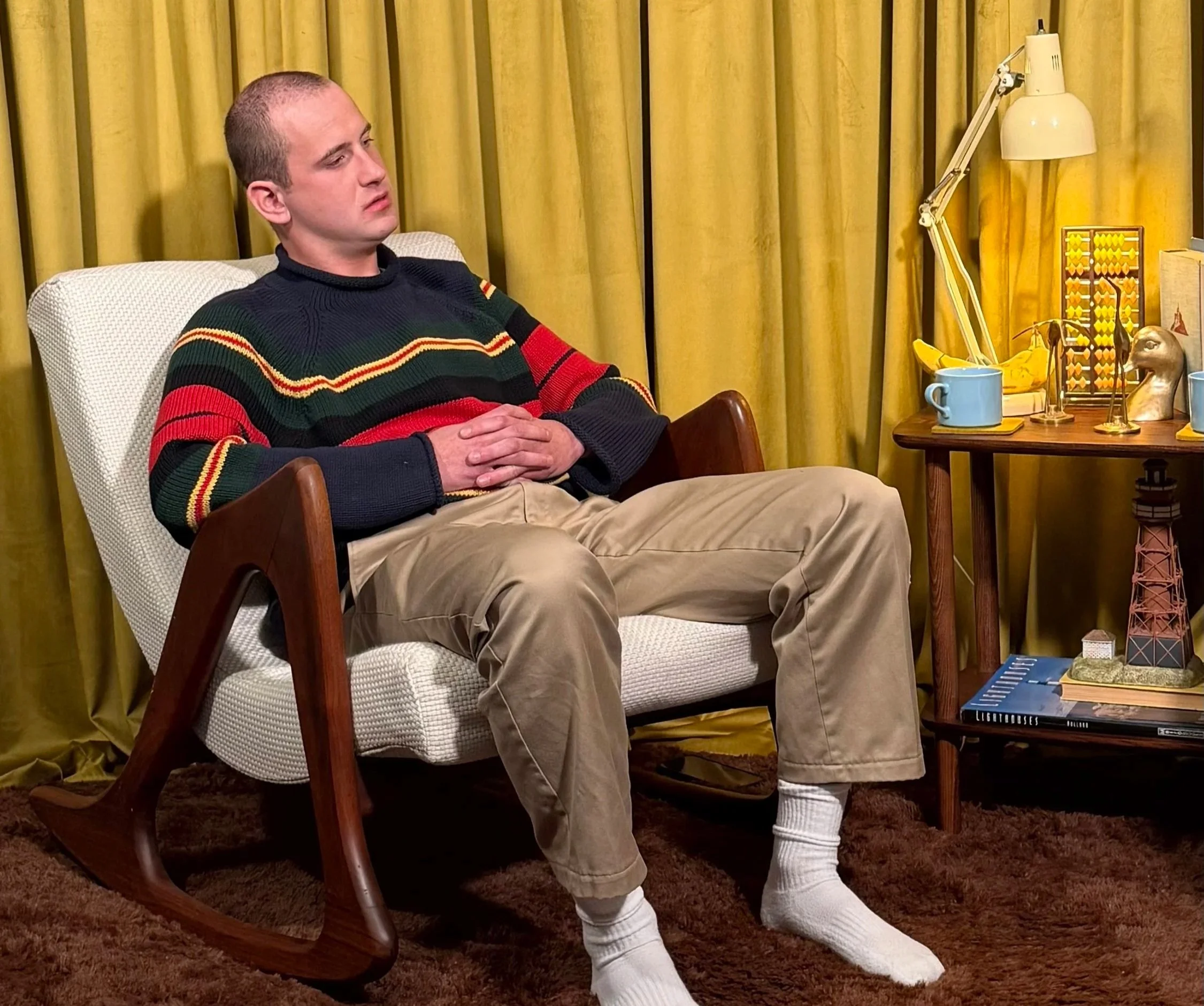 A man sits in a white armchair with his hands clasped, wearing a colorful striped sweater and beige pants, with yellow curtains in the background. A small wooden table beside him holds a blue mug, a lamp, some books, and decorative items.