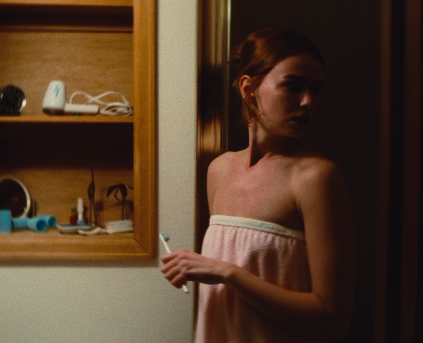 A woman with short red hair wrapped in a pink towel, standing in a dimly lit room, partially visible through a mirror or glass, with a wooden cabinet on the left containing various small objects and toiletries.