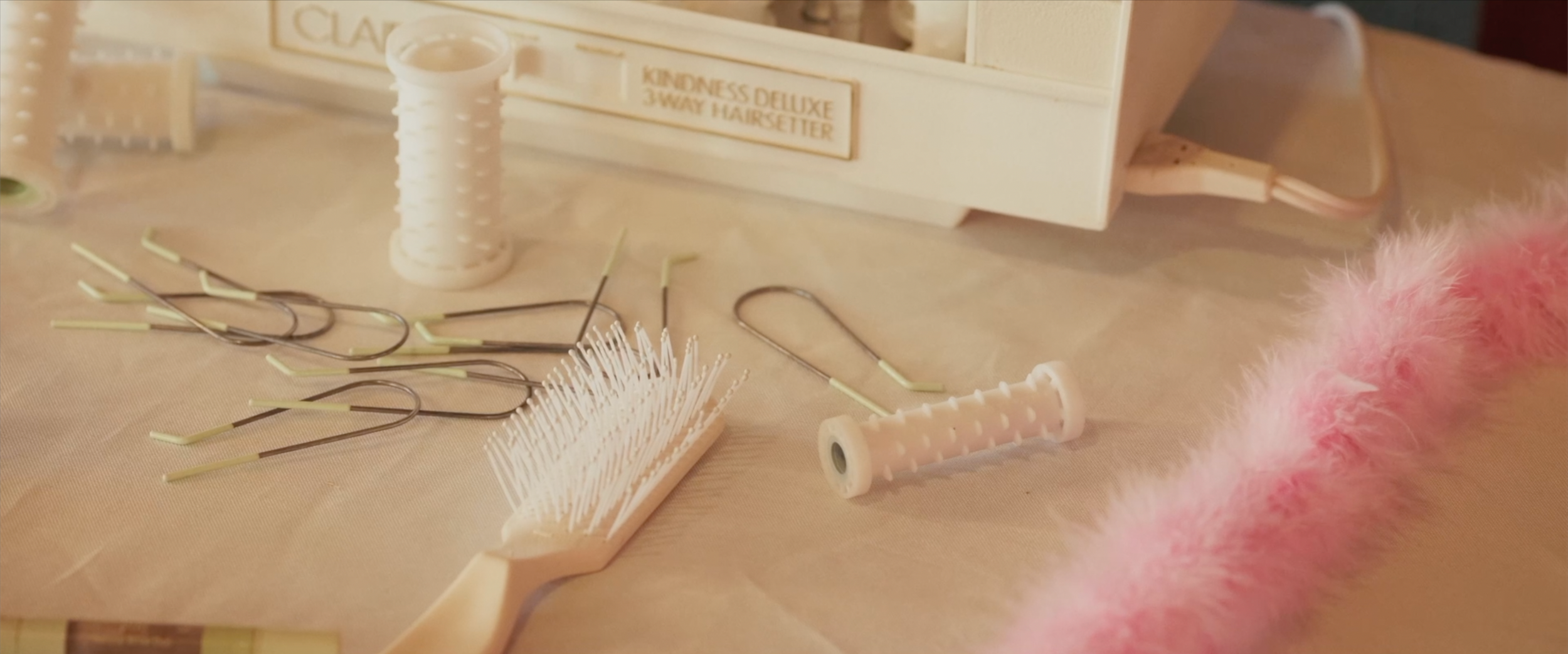 A collection of hair styling tools and accessories, including hairpins, a small round brush, a spool of thread, and a pink fuzzy fabric on a table. In the background, there is a plastic container labeled 'KINDNESS DELUXE 3-WAY HAIRSETTER'.