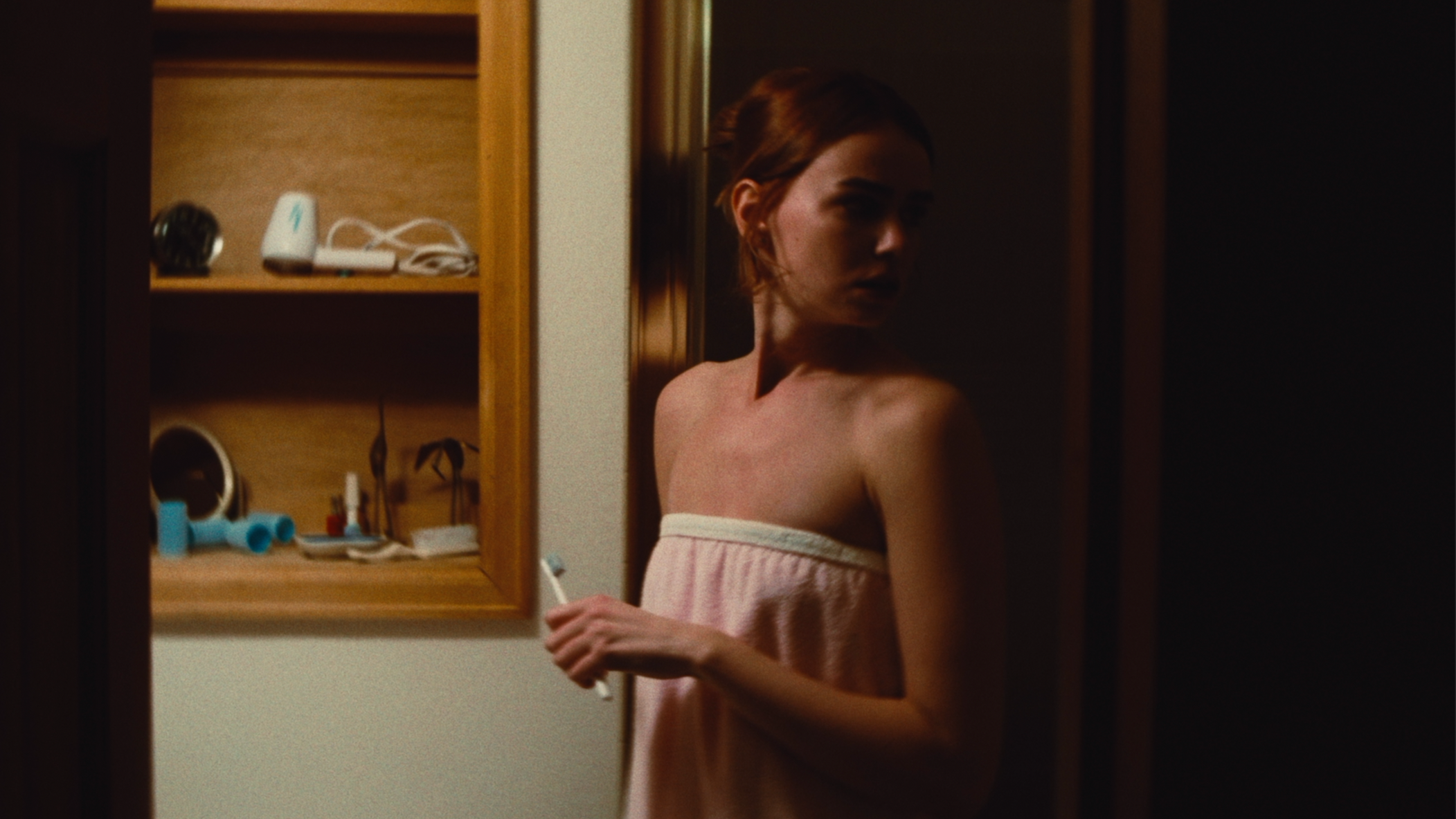 A young woman with short red hair wrapped in a towel, standing in front of a mirror in a bathroom, holding a toothbrush.