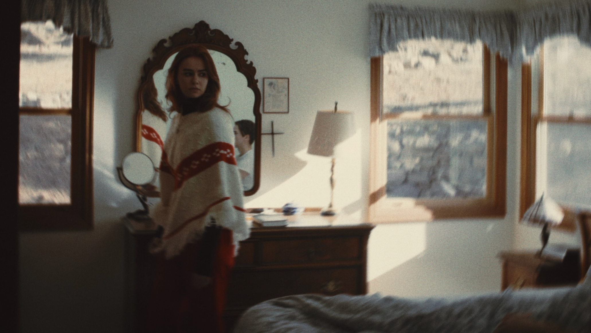 A woman with red hair looking into a mirror in a room with open windows and a view of snow-covered rooftops.