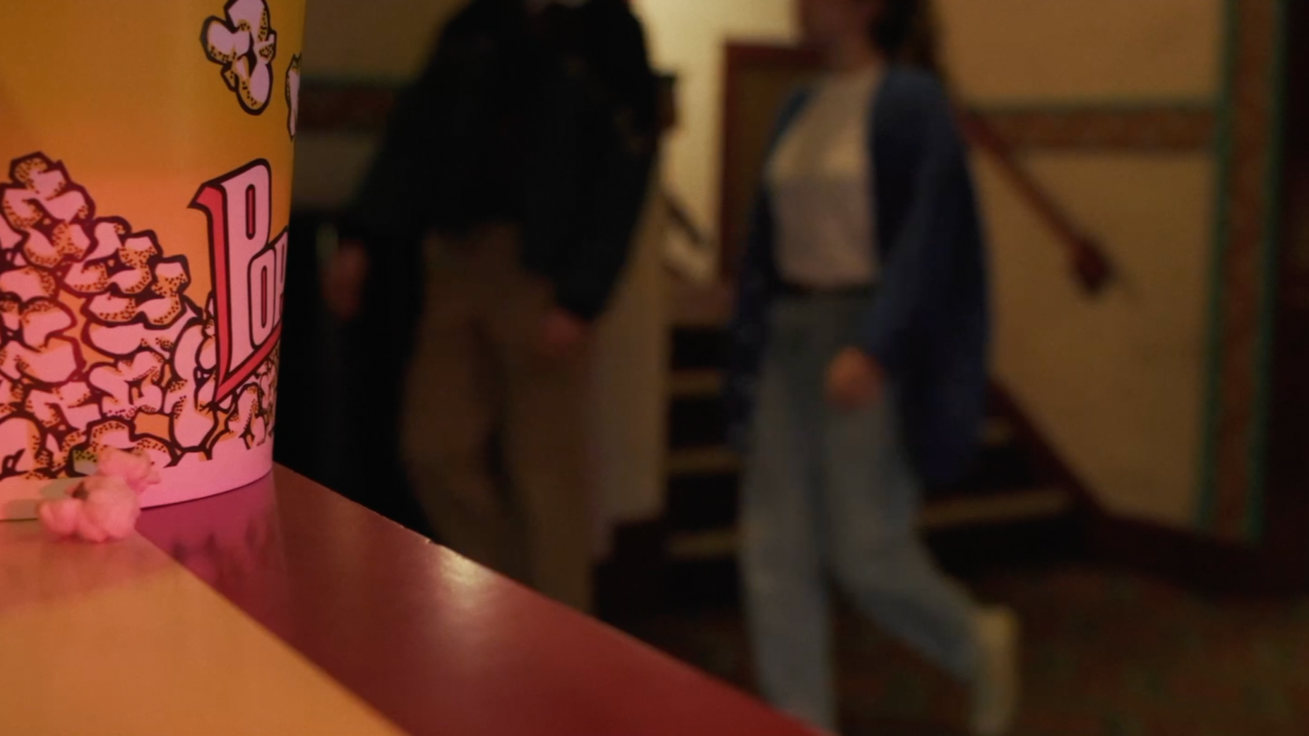 Close-up of a popcorn container with pink and yellow colors on a table. In the background, two people are holding hands near a staircase, but their faces are blurred.