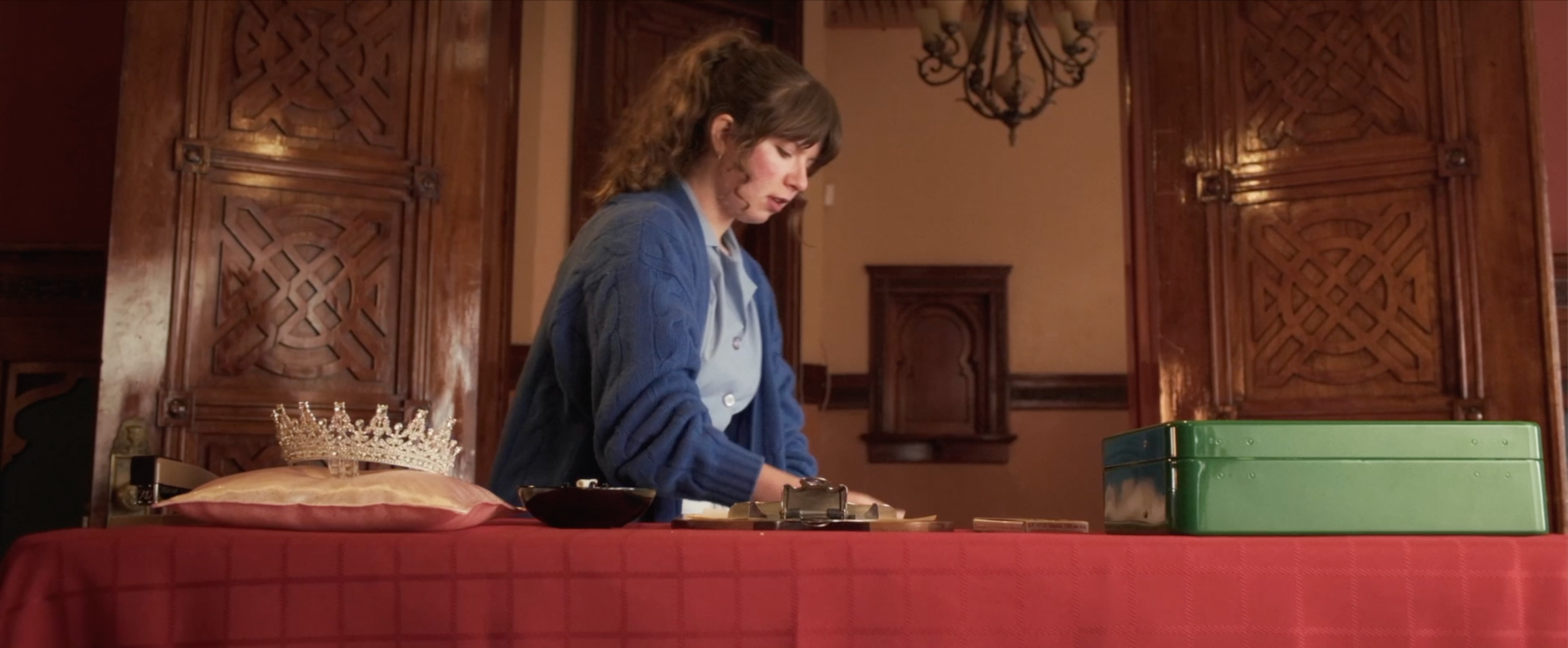 A young woman with brown hair wearing a blue sweater standing at a table with a red tablecloth, organizing items. There is a green metal box, a black bowl, and a small crown resting on a pillow on the table. The room features wood-paneled walls, orna
