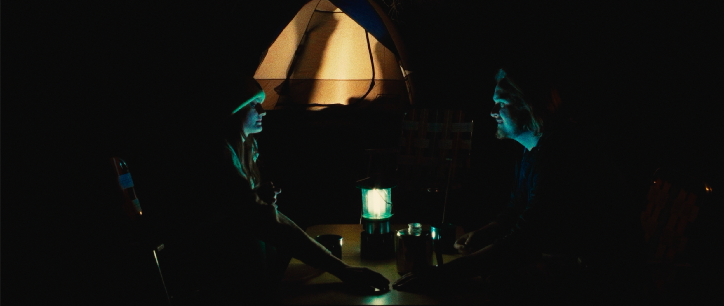 Two people sitting at a table in the dark, illuminated by a lantern, with a tent in the background.