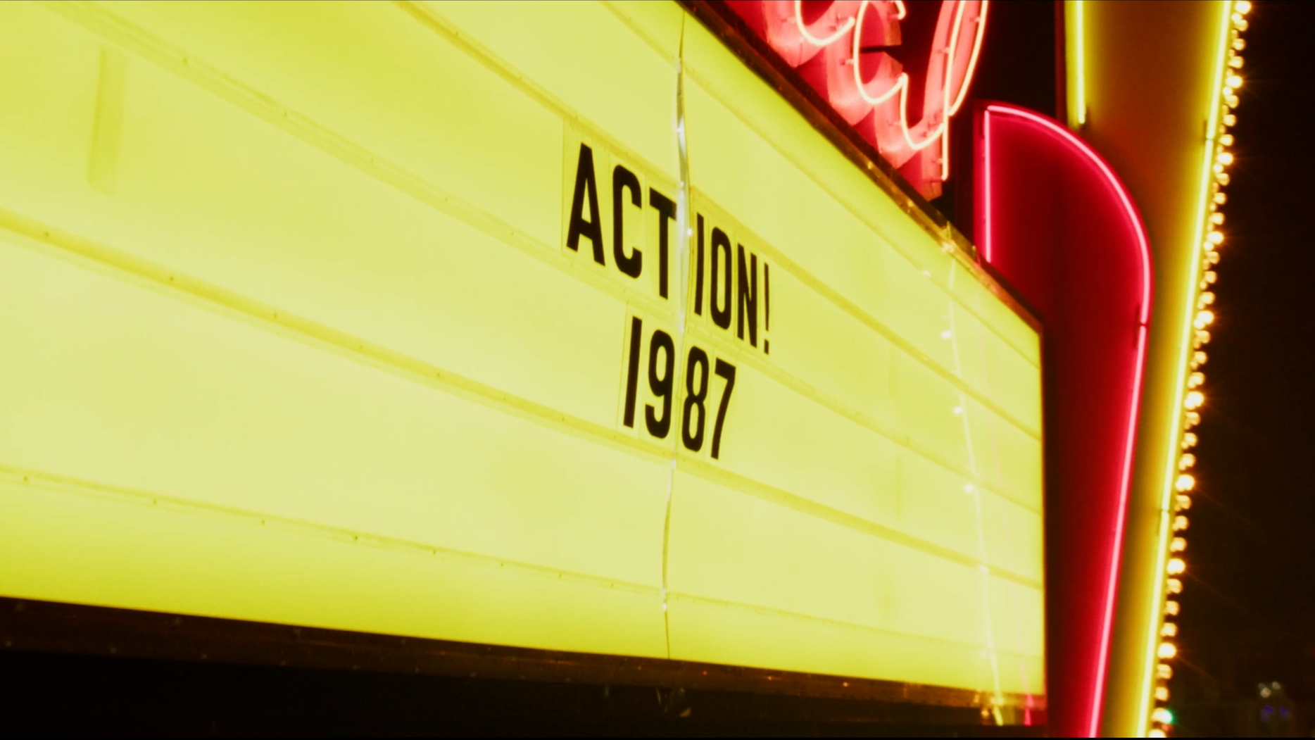 A neon-lit yellow marquee sign with black lettering stating 'ACTION! 1987', partially visible neon lights in red and yellow at the top.