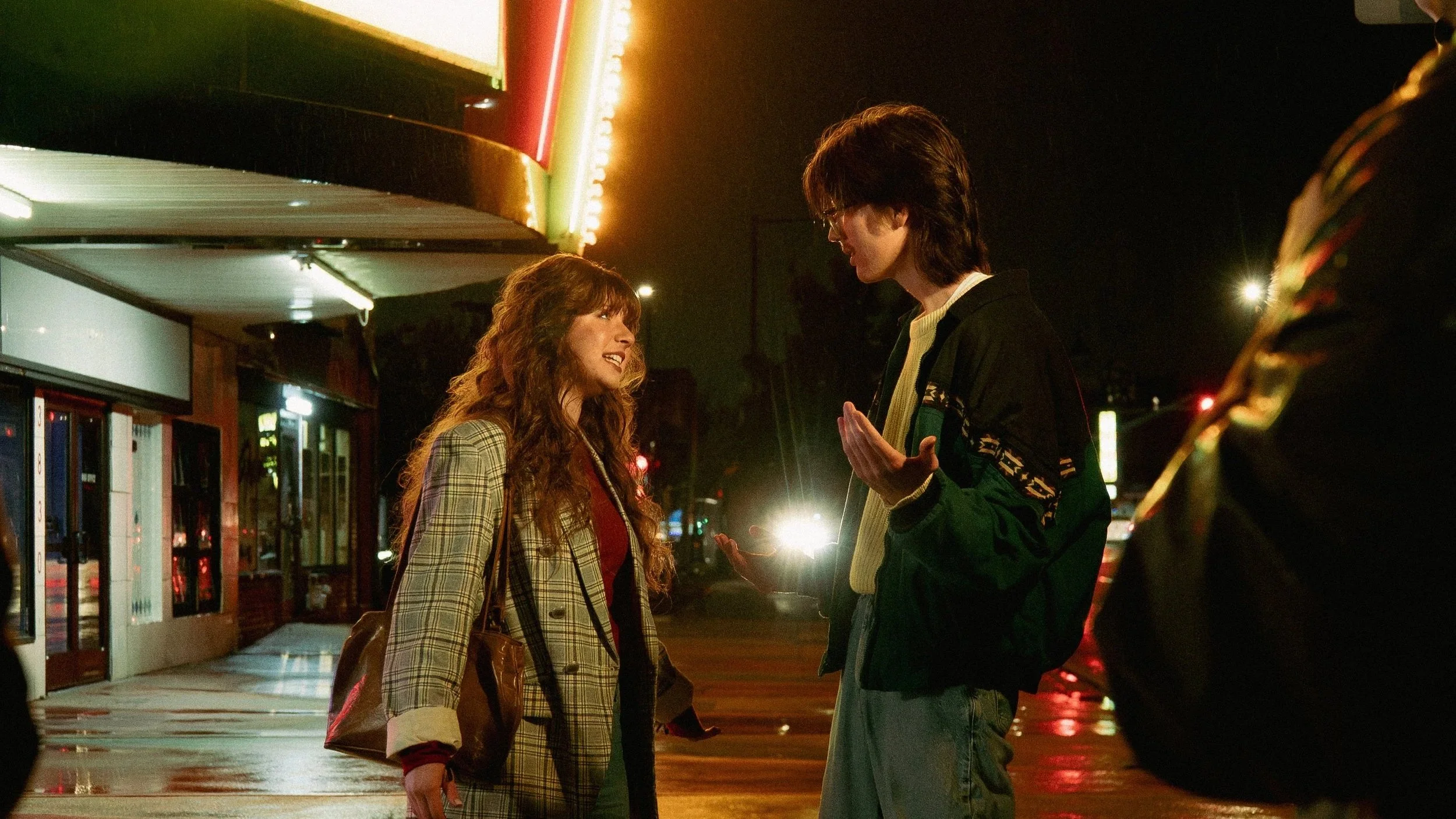 A man and woman talking on a nighttime street with neon lights, sidewalk, and a vending machine visible.