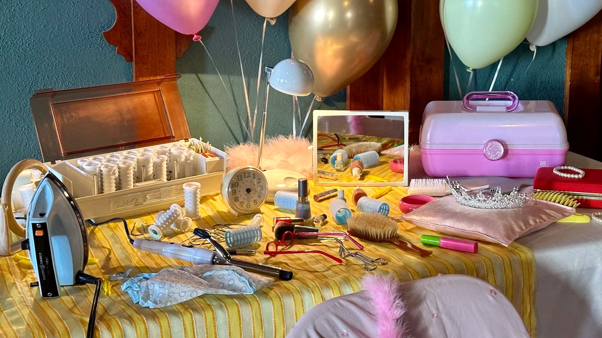 A table covered with a yellow-striped tablecloth holding various cosmetic and beauty items, including a white plastic storage case with nail polish bottles, an iron, a clock, a mirror, hairbrushes, bottles, scissors, curlers, a tiara, and a pink cush