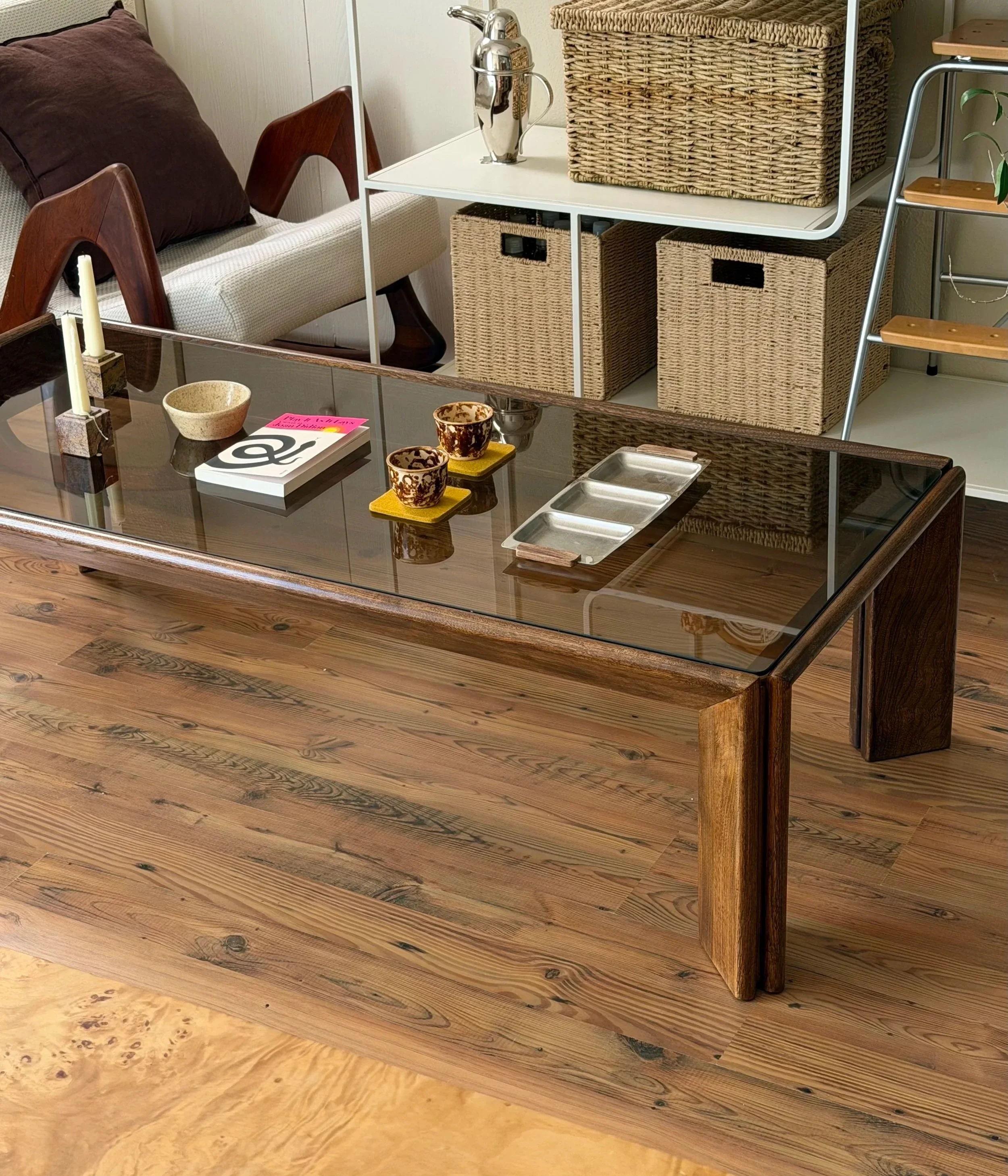 Living room area with a wooden coffee table, woven baskets on a white metal shelving unit, a ceramic vase, a beige upholstered sofa with a brown pillow, and a wooden and metal stairs leading to an upper level. The floor is hardwood.