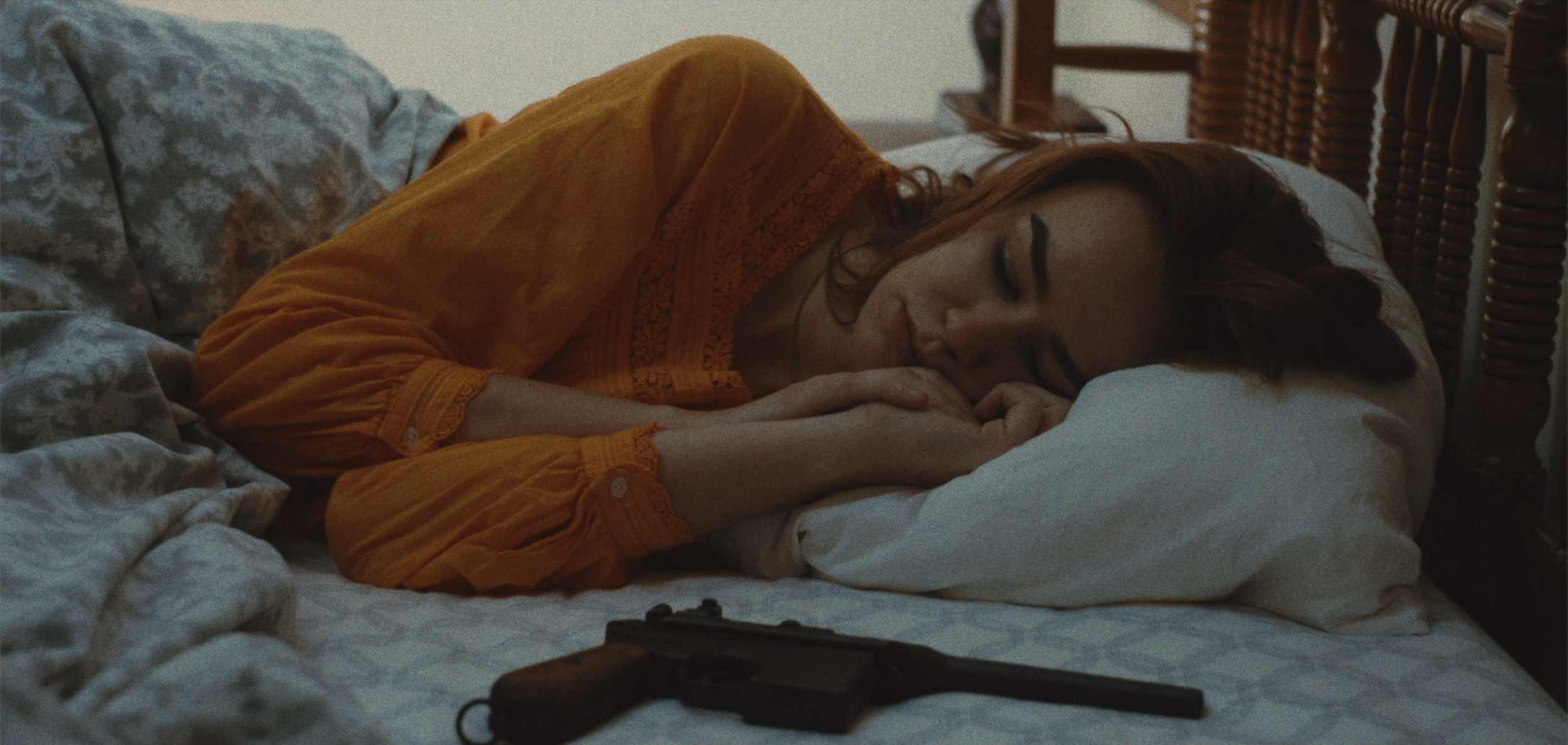 A woman with red hair sleeping on a bed, wearing an orange blouse, with a handgun placed on the bed in front of her.