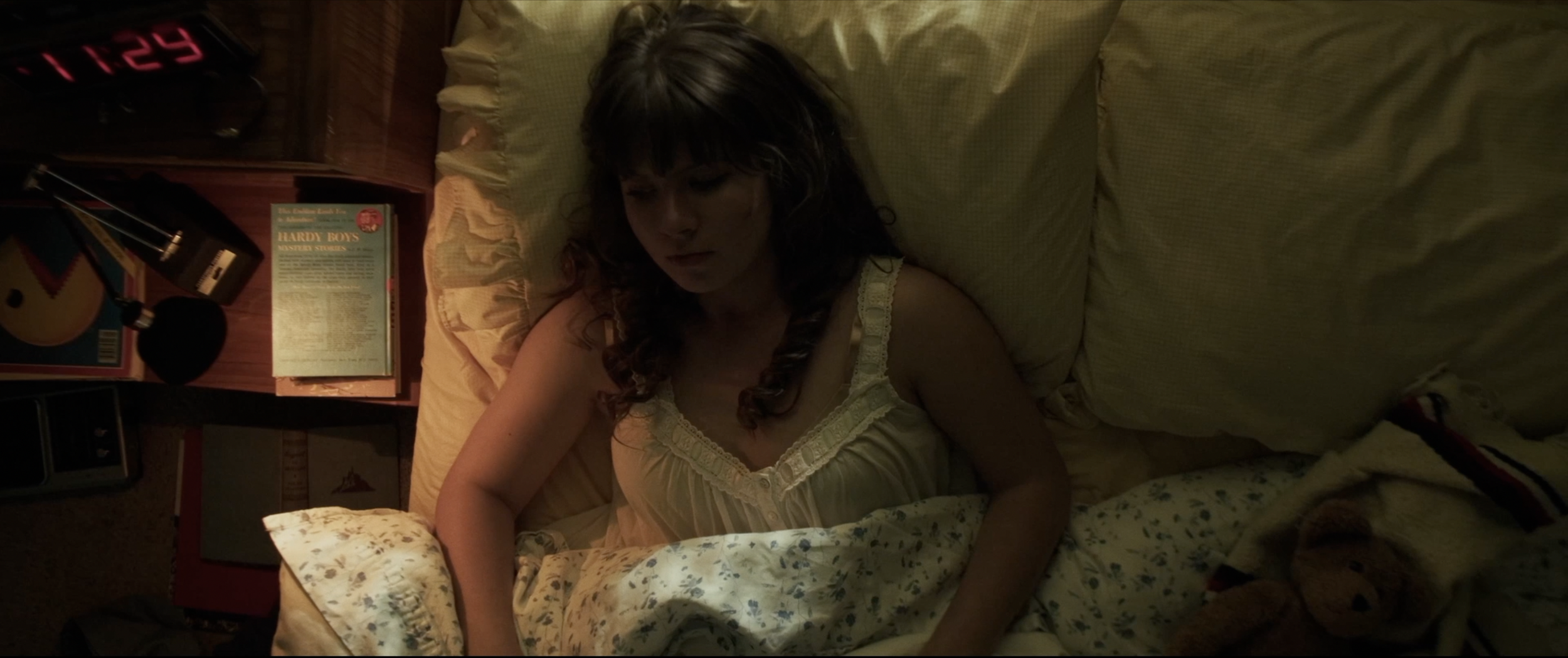A woman with wavy brown hair sleeping in a bed with yellowish bedding, surrounded by a nightstand with books, a clock, and other items in a dimly lit room.
