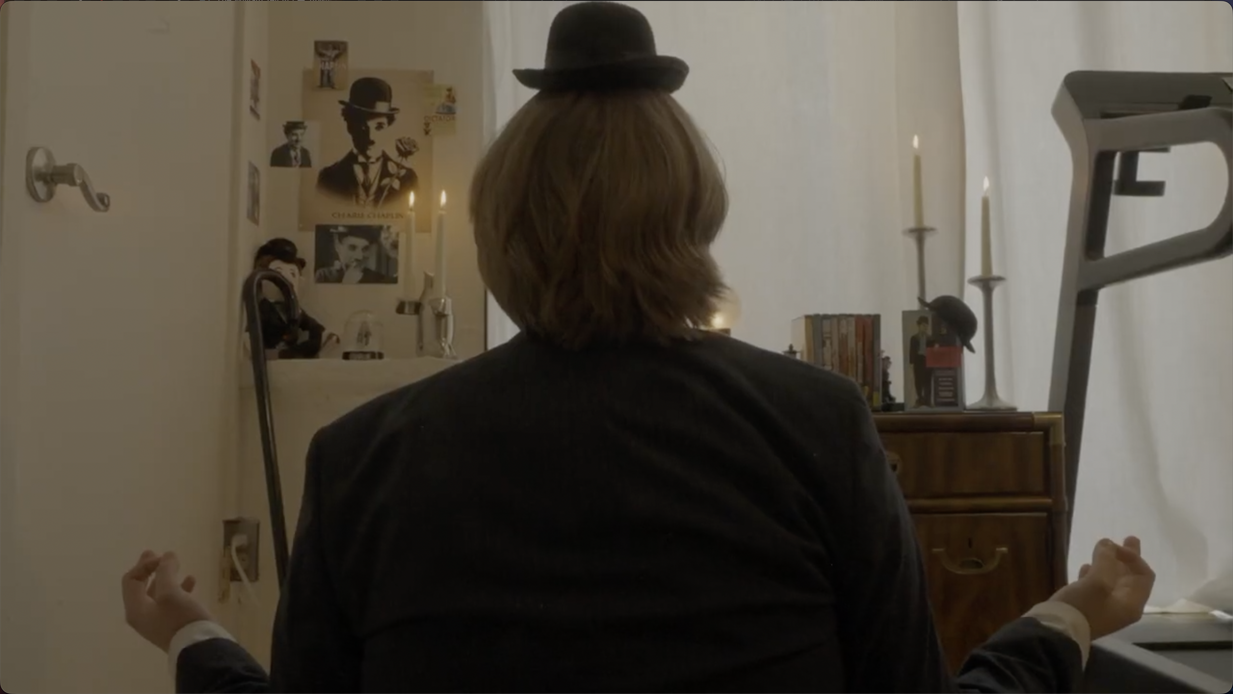 Person with shoulder-length hair and a hat, sitting with hands in a meditative pose in front of a desk with books and candles, in a room decorated with Cheristmas posters on the wall.