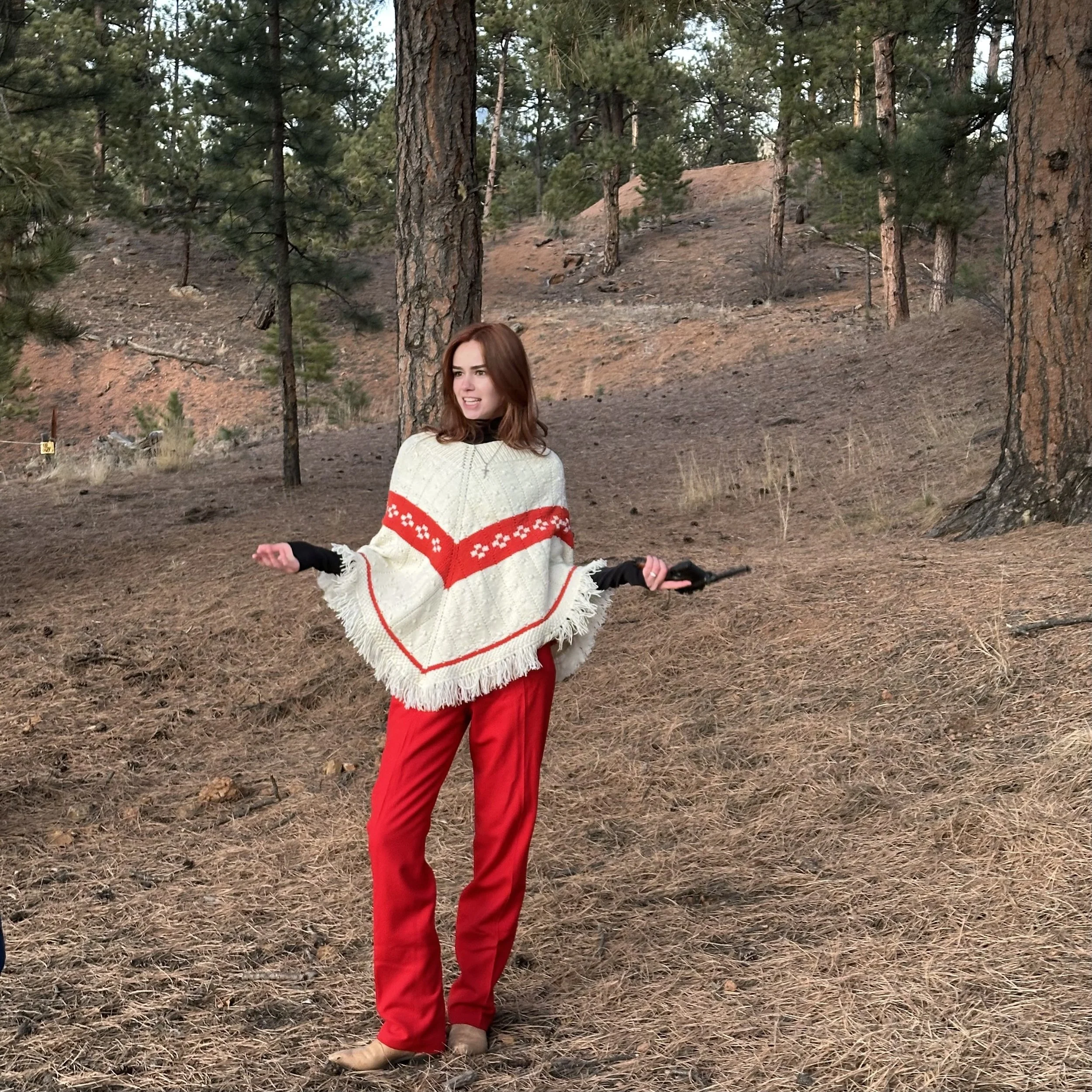 A woman with long brown hair stands outdoors in a forested area with pine trees, wearing a cream-colored sweater with red and black accents and red pants.
