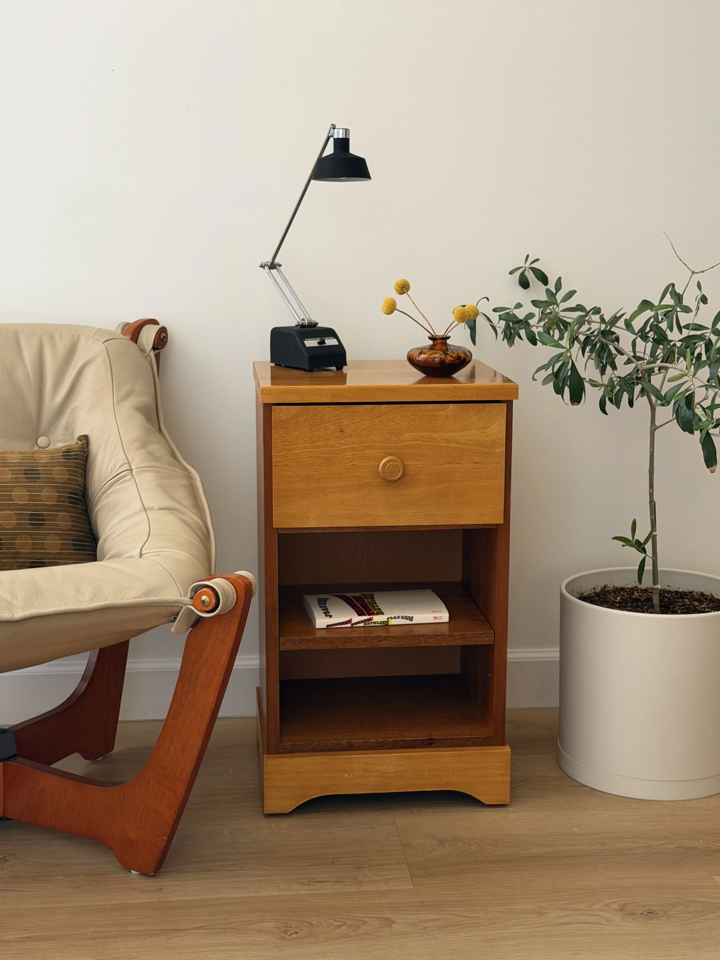 A beige armchair with a patterned pillow on the left, a wooden side table with a black adjustable desk lamp, a brown vase with yellow flowers, a book on the bottom shelf, and a large potted plant on the right, set against a white wall and wooden floo