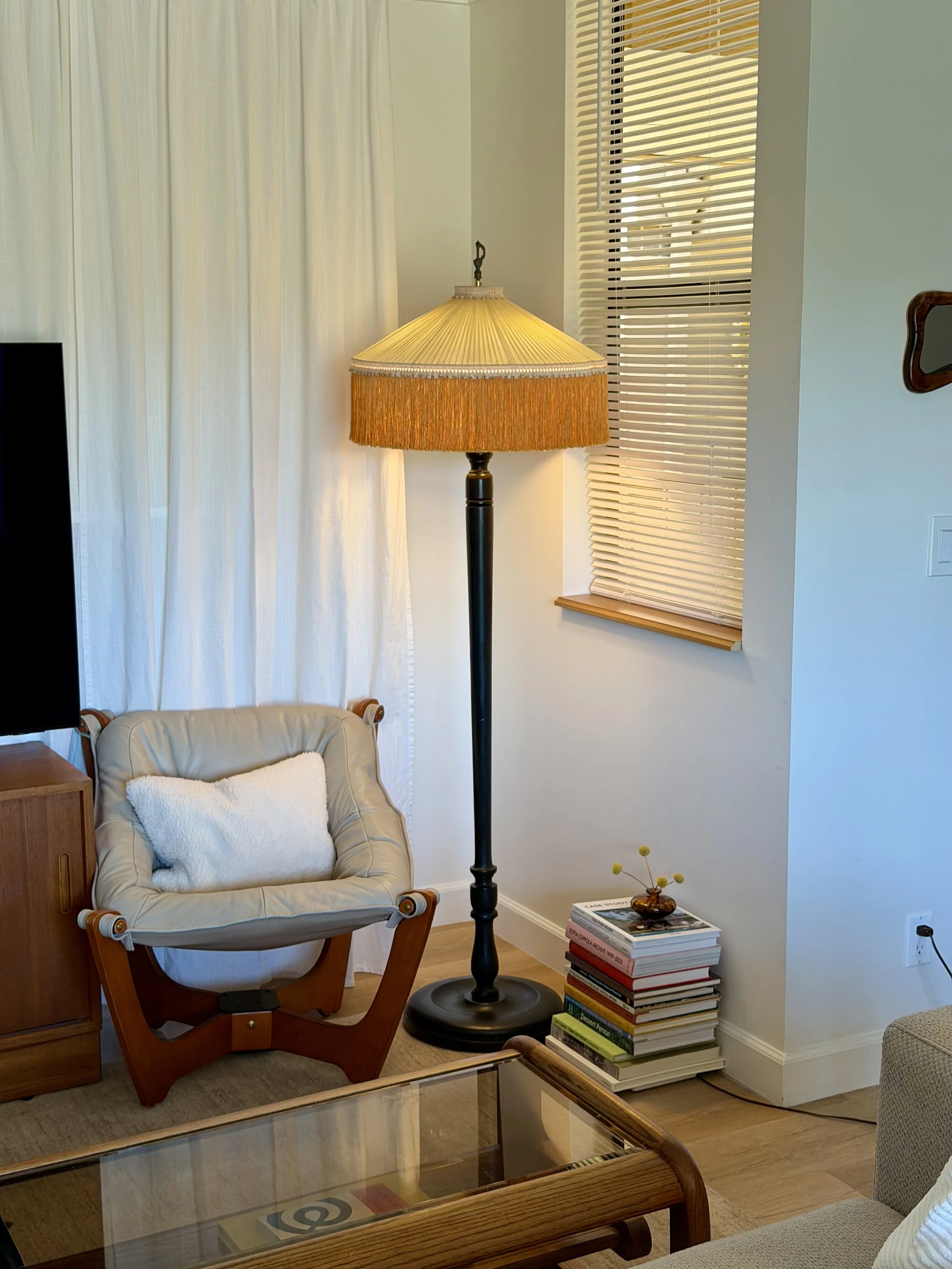 A cozy living room corner with a beige armchair, a tall floor lamp with a fringed lampshade, a small stack of books topped with a decorative flower arrangement, and a glass coffee table.