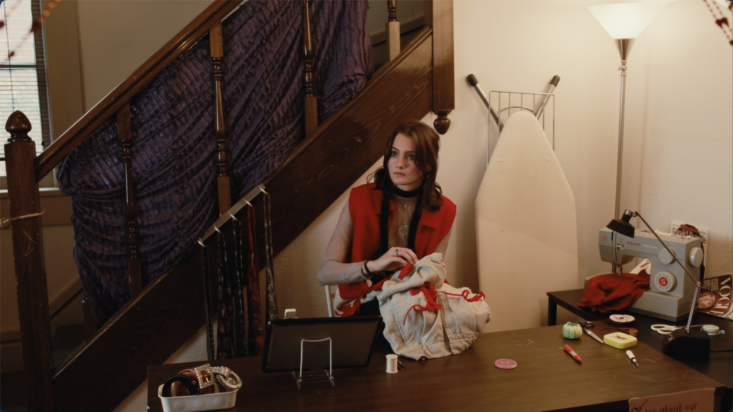 A young woman with brown hair and pale skin sitting at a sewing station under a staircase. She is holding a piece of clothing and looking attentively to her right. The workspace includes a Singer sewing machine, fabric, scissors, pins, and other sewi