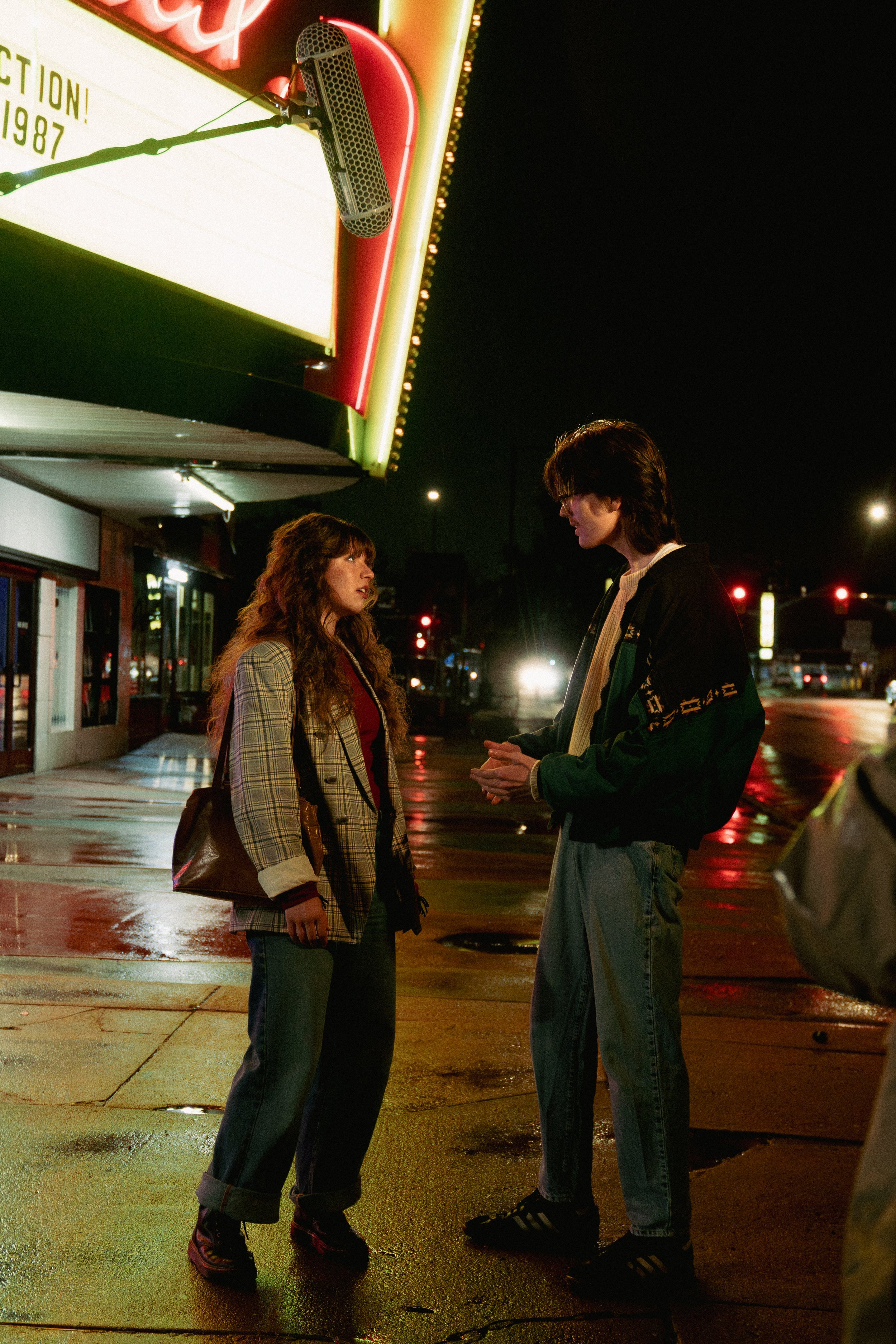 Two young adults, a woman with curly hair and a man with straight hair and glasses, stand on a wet city sidewalk at night, engaged in conversation under the glow of neon signs. The woman wears a plaid blazer and dark pants, and the man wears a dark j