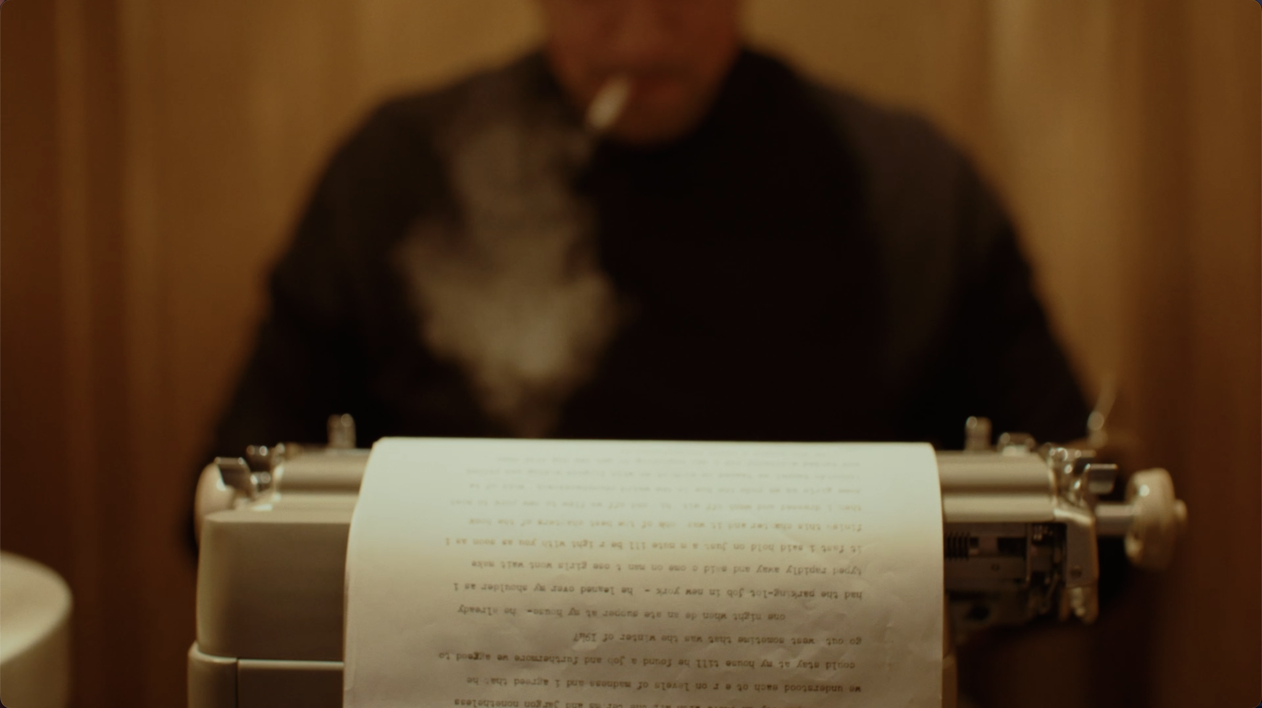 A person typing on a typewriter with a sheet of paper inserted, smoke rising from a cigarette in their mouth, in a warm-colored room.