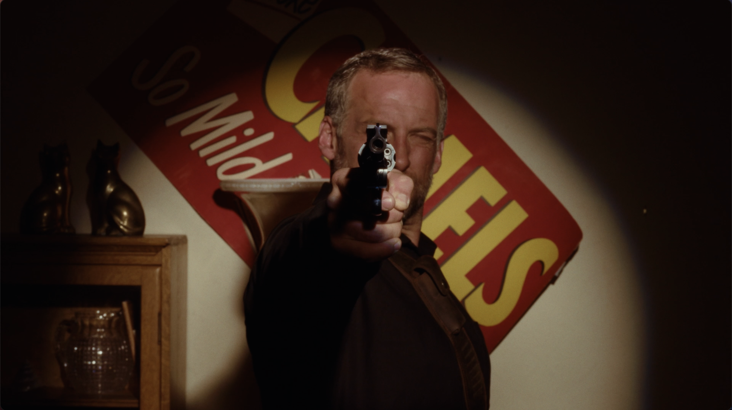 A man pointing a handgun directly at the camera, with a large colorful sign in the background, and a wooden cabinet with decorative items to the left.