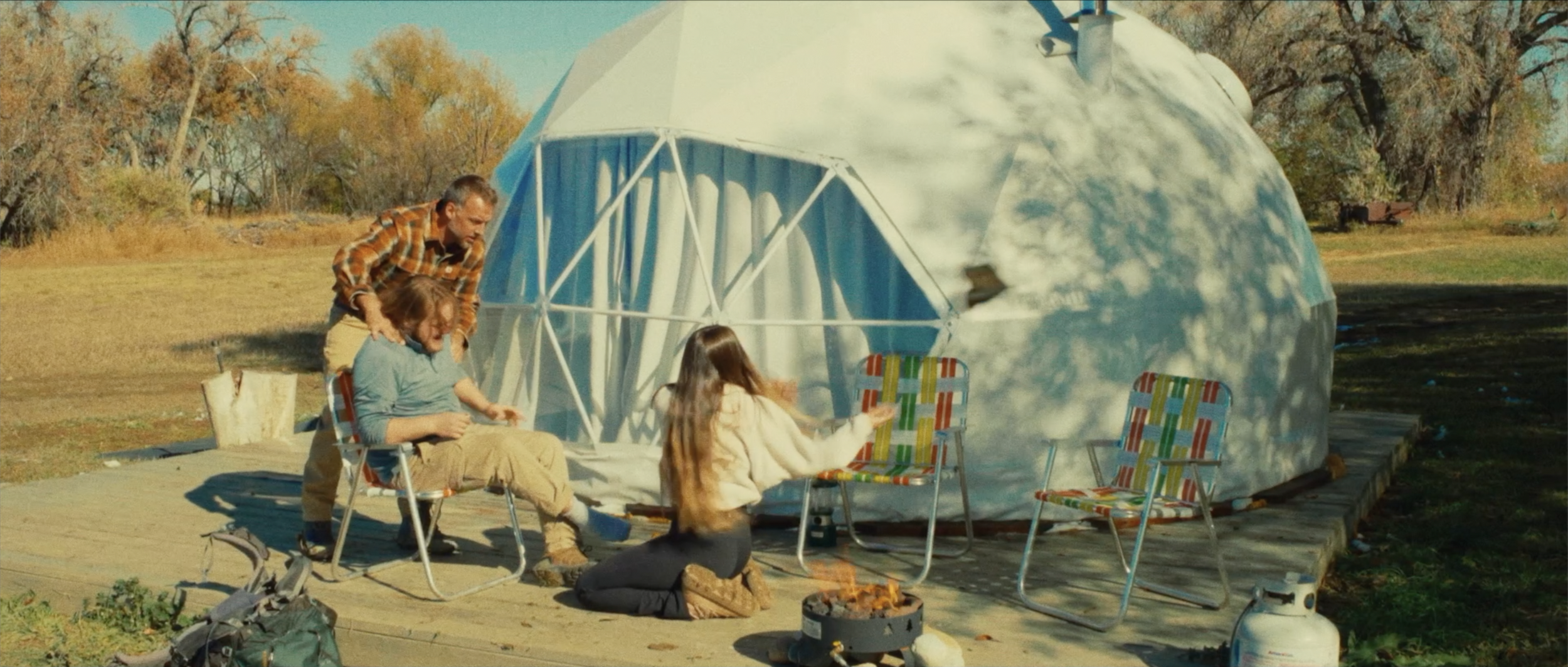 People sitting outside in front of a geodesic dome, with wood chairs, a small fire pit, and camping gear, during autumn with trees in the background.