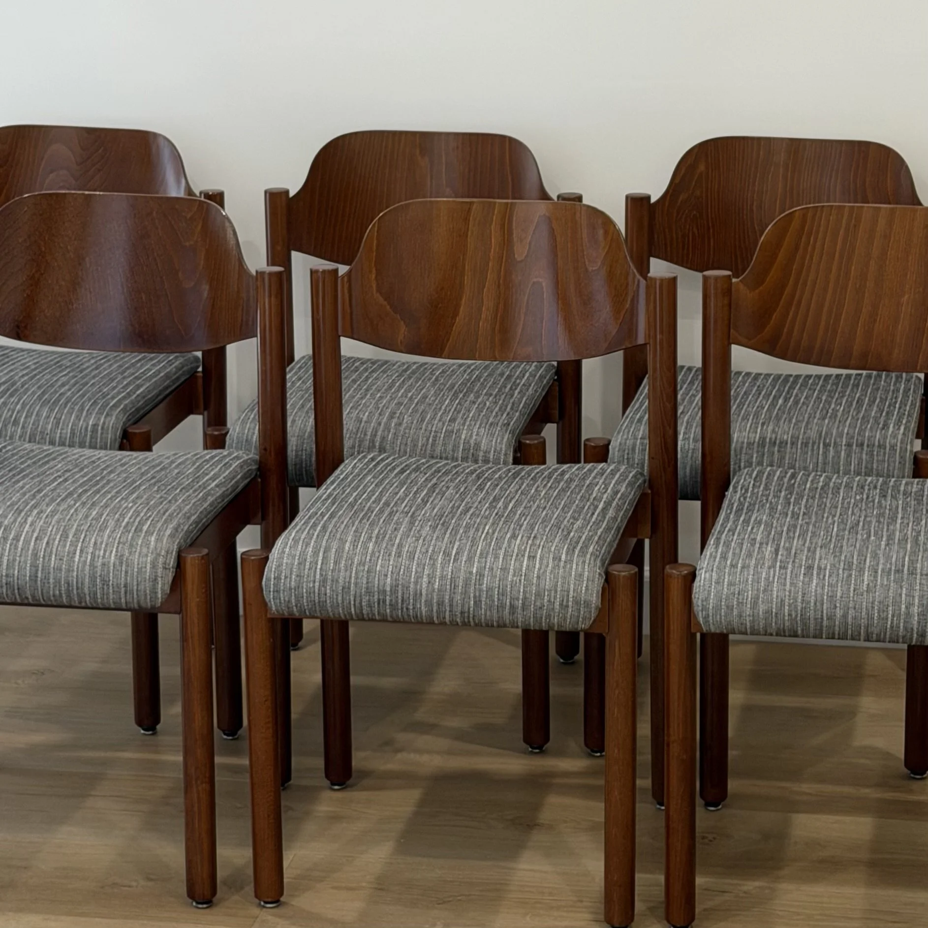 Six wooden chairs with dark striped fabric seats arranged in two rows on a wooden floor against a plain light-colored wall.
