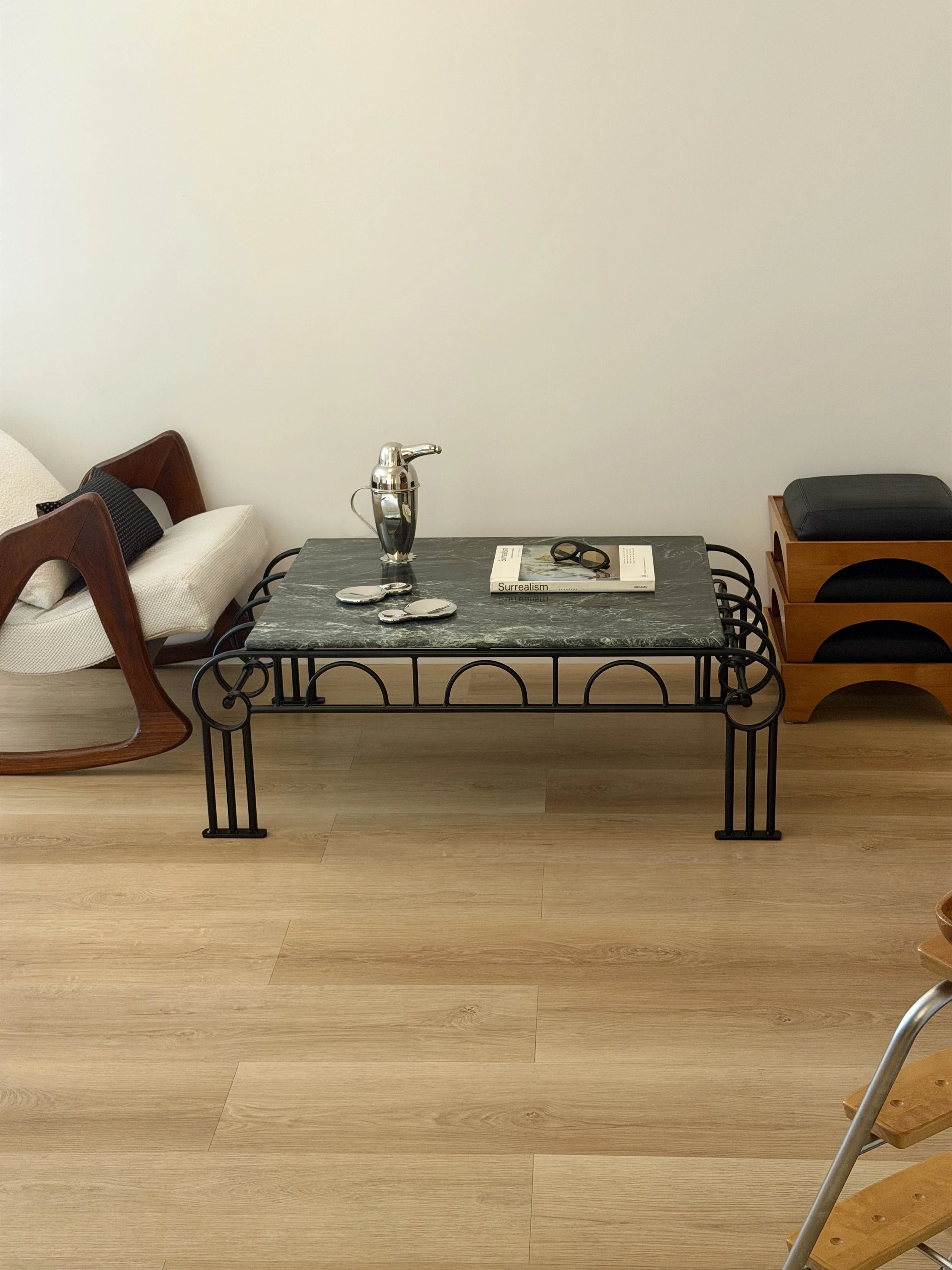 A minimalist interior scene featuring a black metal coffee table with a gray marble top, a book titled "Surrealism" and a pair of black glasses on it. To the left, there is a vintage wooden chair with a curved backrest, and on the right, a wooden cab