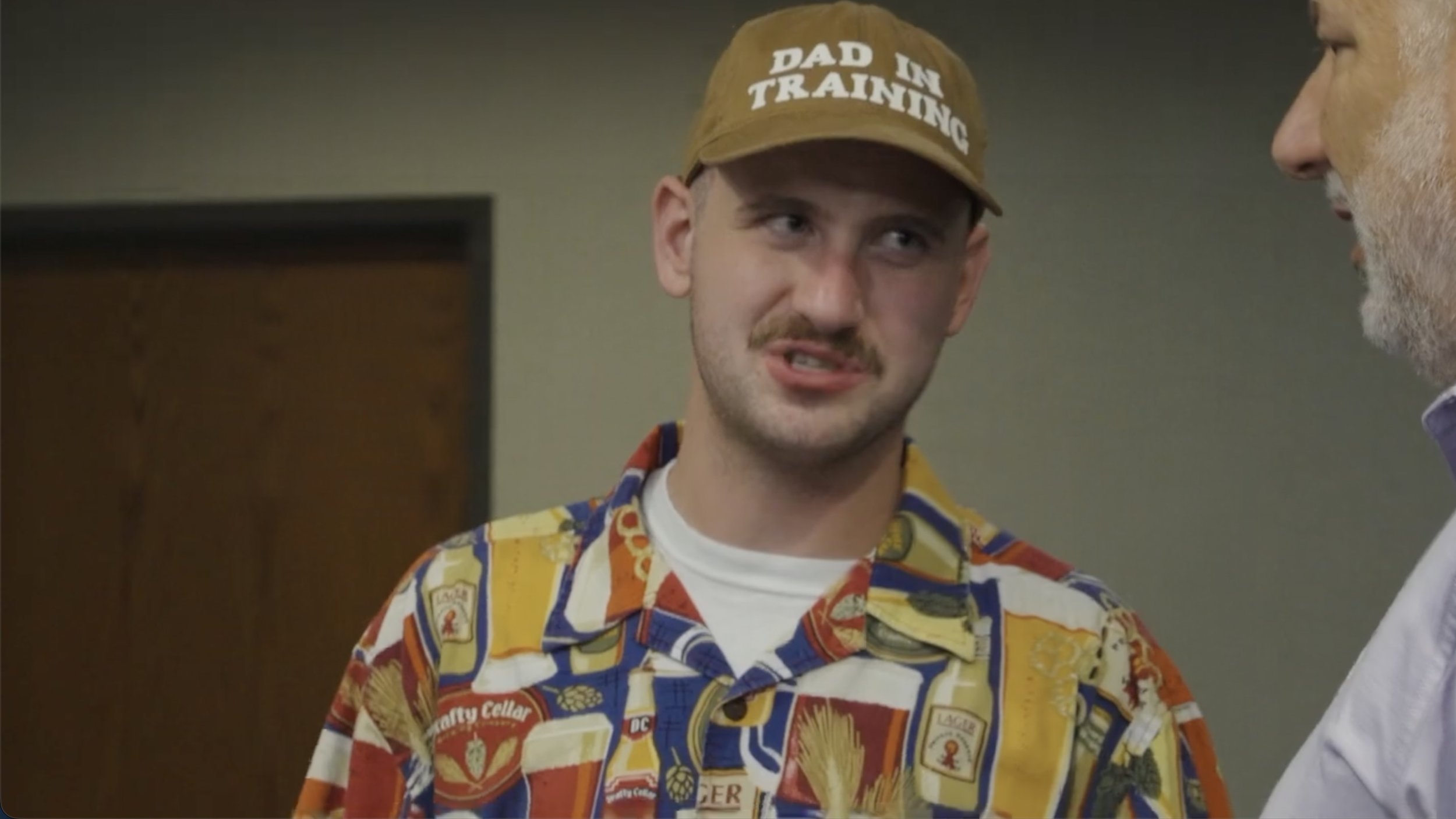 A young man wearing a colorful Hawaiian shirt and a tan cap that says 'Dad Line Training' is talking to an older man with white hair and a beard.