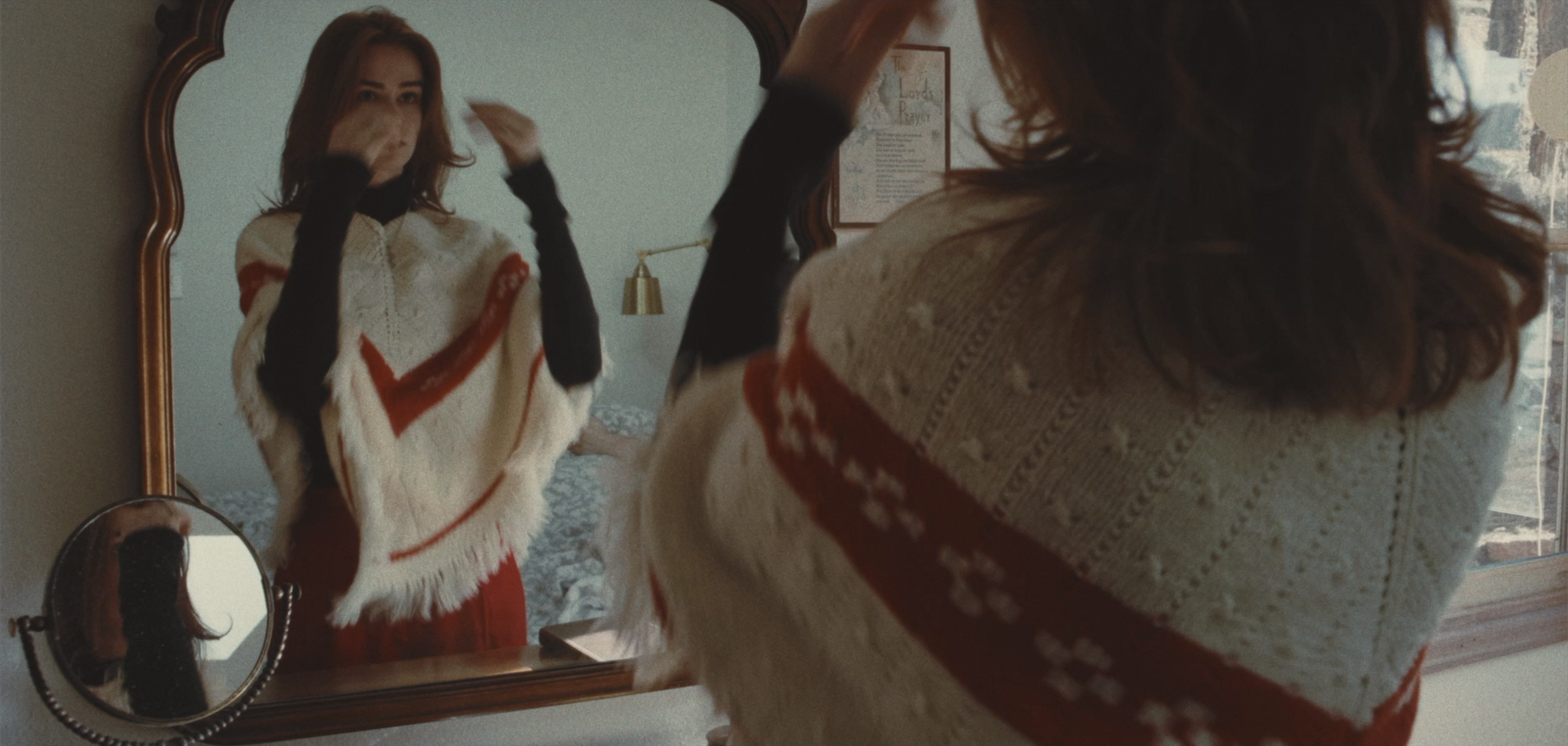 A woman looking at herself in a mirror, adjusting her hair, wearing a Christmas sweater with red and white patterns.