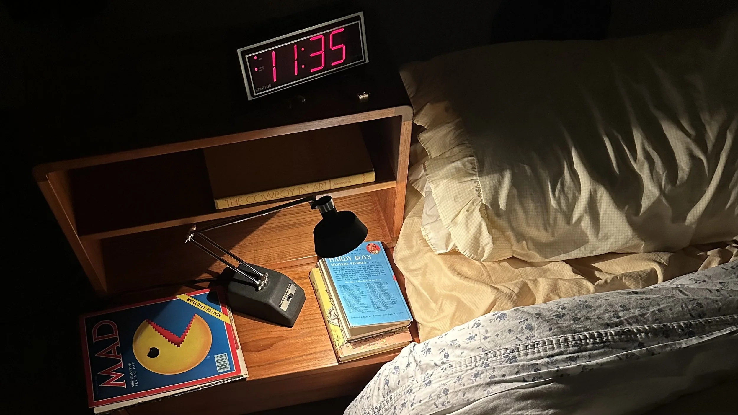 A bedside table with a black lamp, several books including 'The Cowboy in Art,' 'Hardy Boys Mystery Stories,' and 'Mandate for the 21st Century,' a phone charger, and a vintage alarm clock displaying 11:35 p.m. The bed has beige and floral patterned 