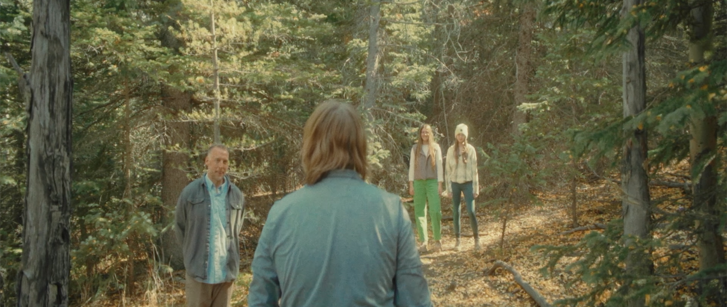 A person with shoulder-length brown hair, wearing a blue jacket, facing away from the camera, standing in a forest. Four people, including a man and three women, are standing in front of this person, among trees with green and yellow leaves, indicati