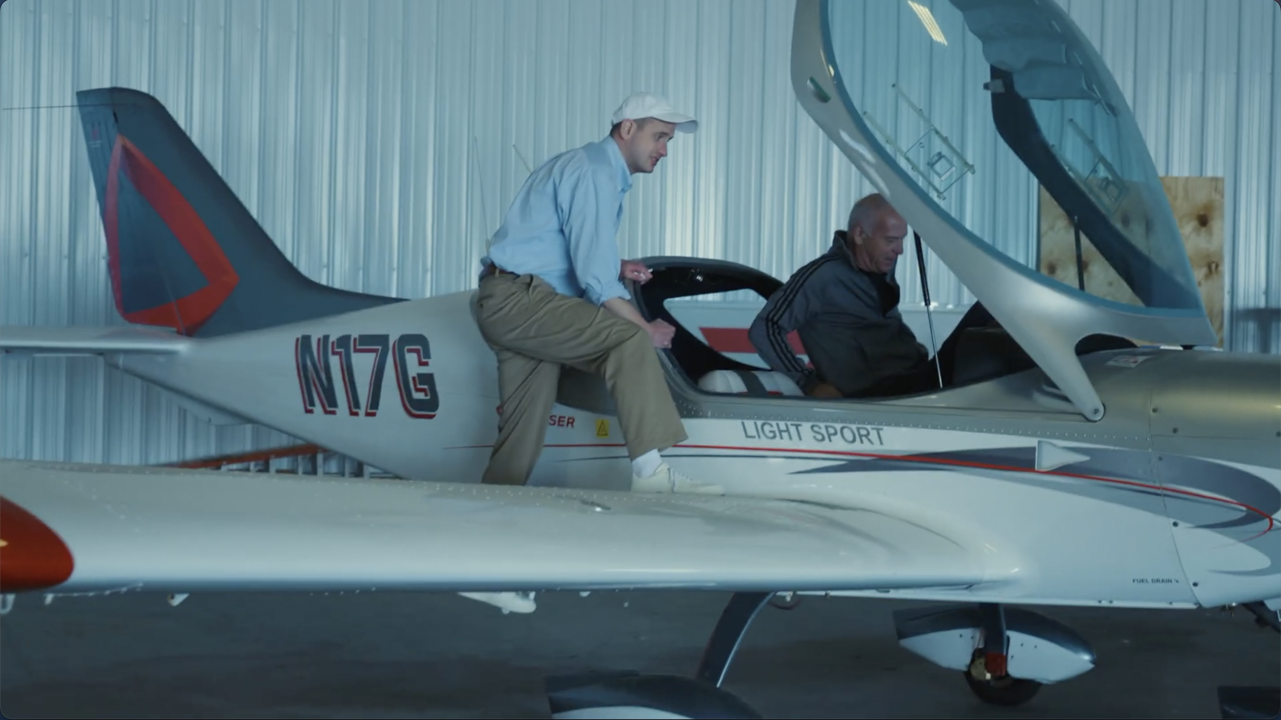 Two men near a small aircraft inside a hangar, one sitting on the edge of the plane's cockpit and the other standing beside him. The aircraft has 