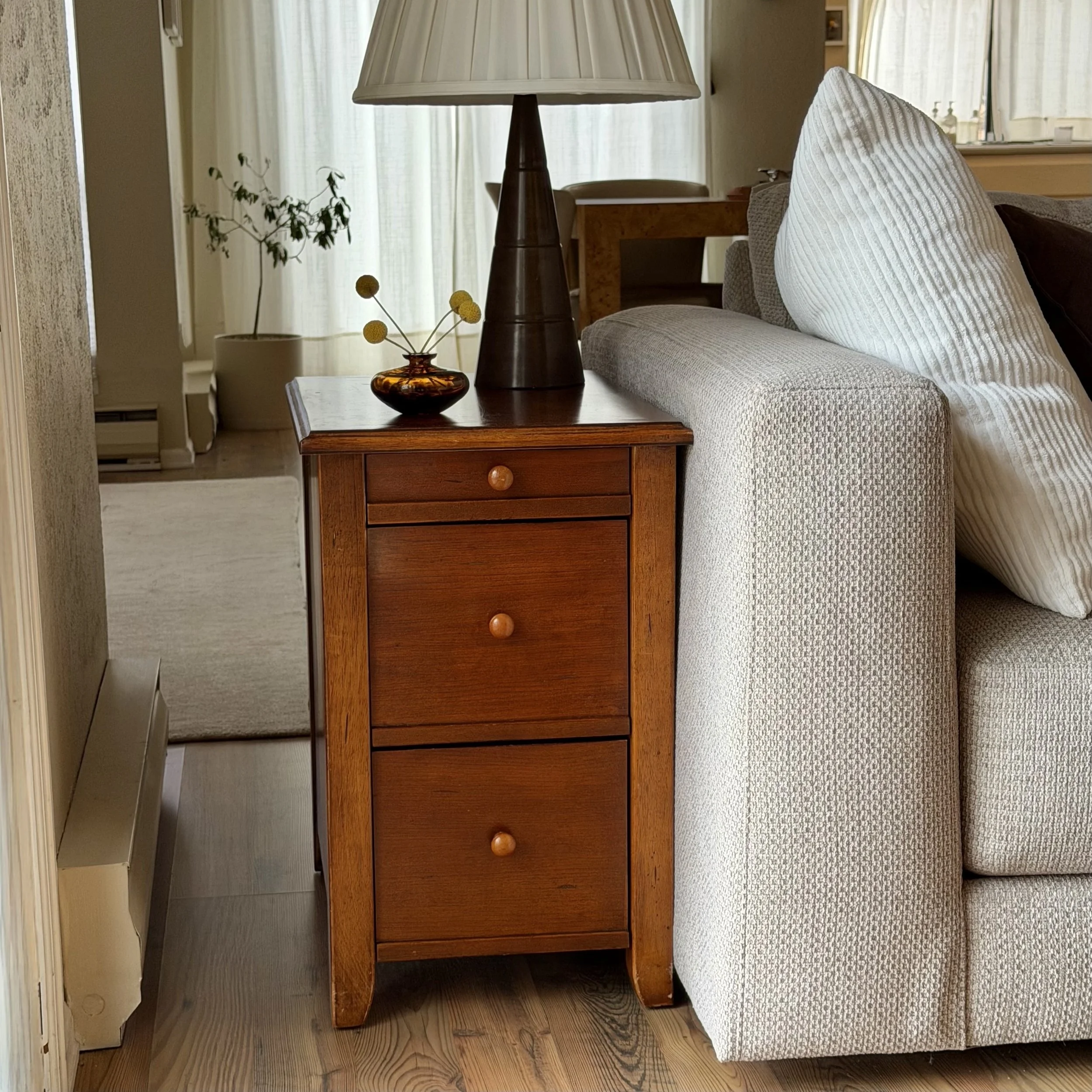 A wooden nightstand with a ceramic vase with yellow spherical flowers, a table lamp with a beige shade, next to a light-colored upholstered sofa with a textured white pillow, in a cozy living room with sheer curtains and hardwood flooring.