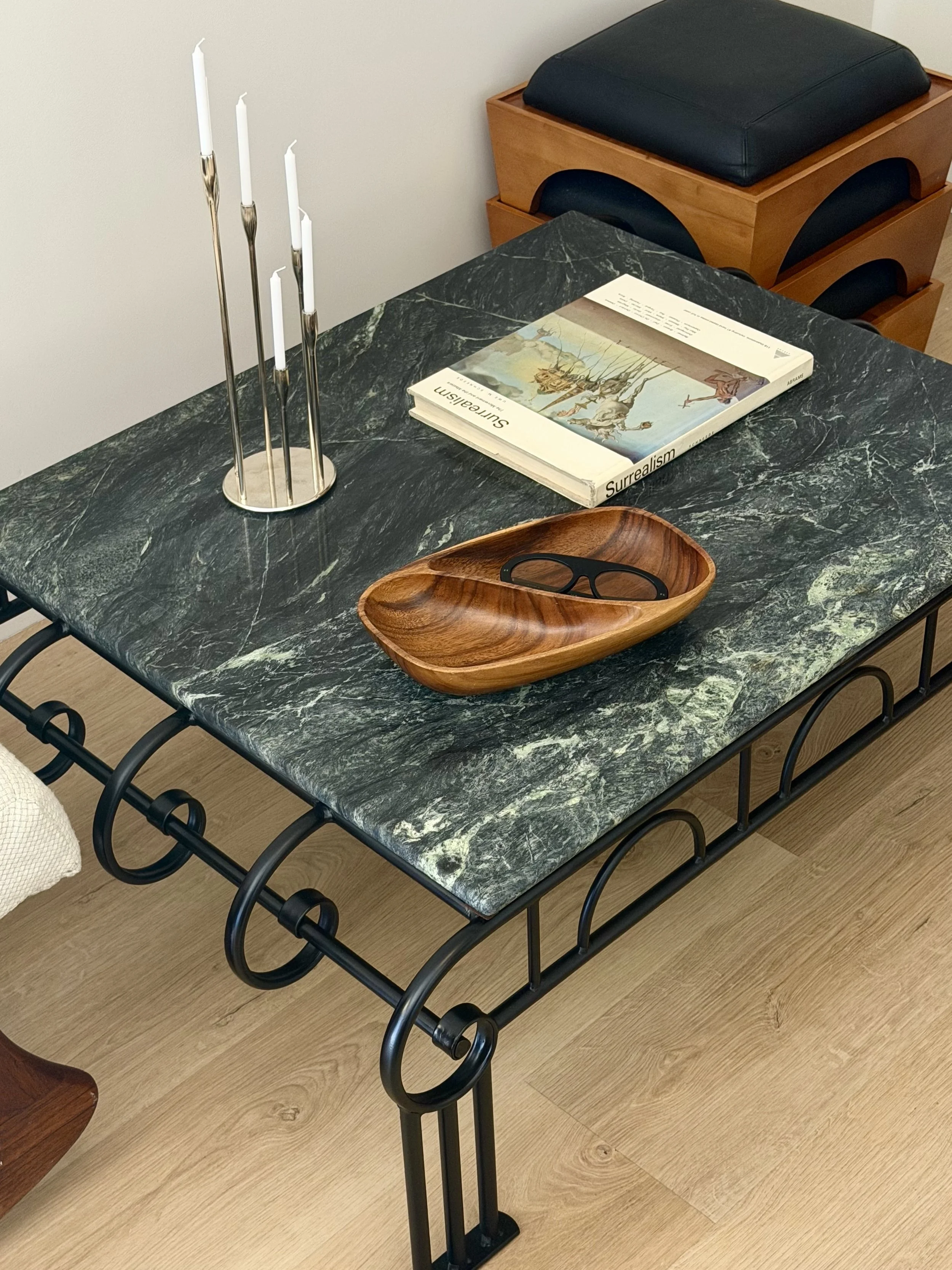 Black marble table with a brass candle holder with five white candles, a paperback book titled 'Surrealism' with artwork on the cover, a small wooden dish with eyeglasses, and wooden furniture with black cushions in the background.