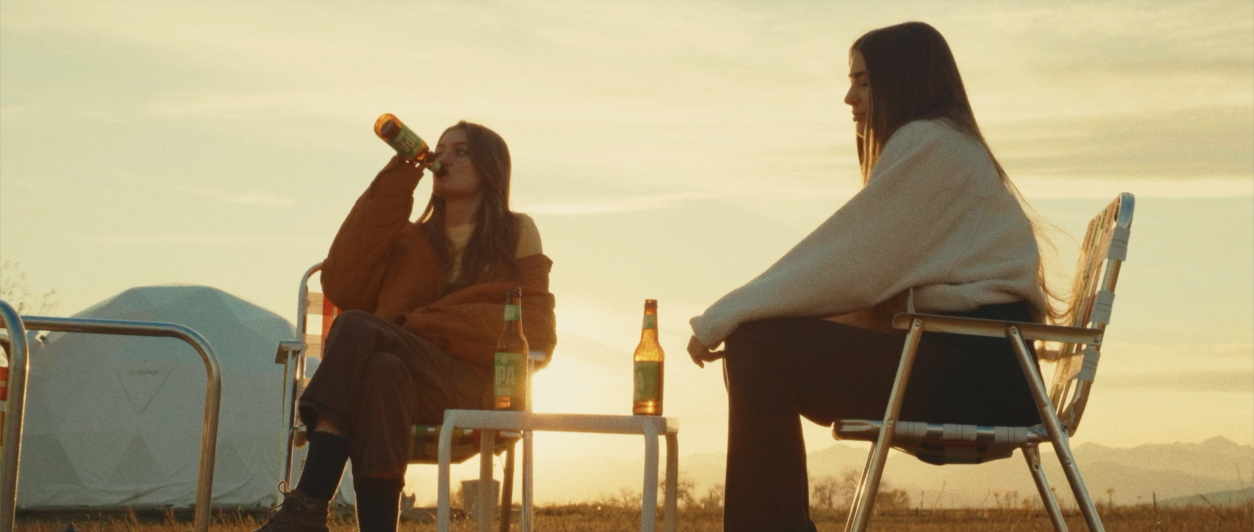 Two women sitting outdoors on camping chairs near a tent during sunset, each with a beer bottle, one woman drinking from her bottle, the other sitting peacefully.