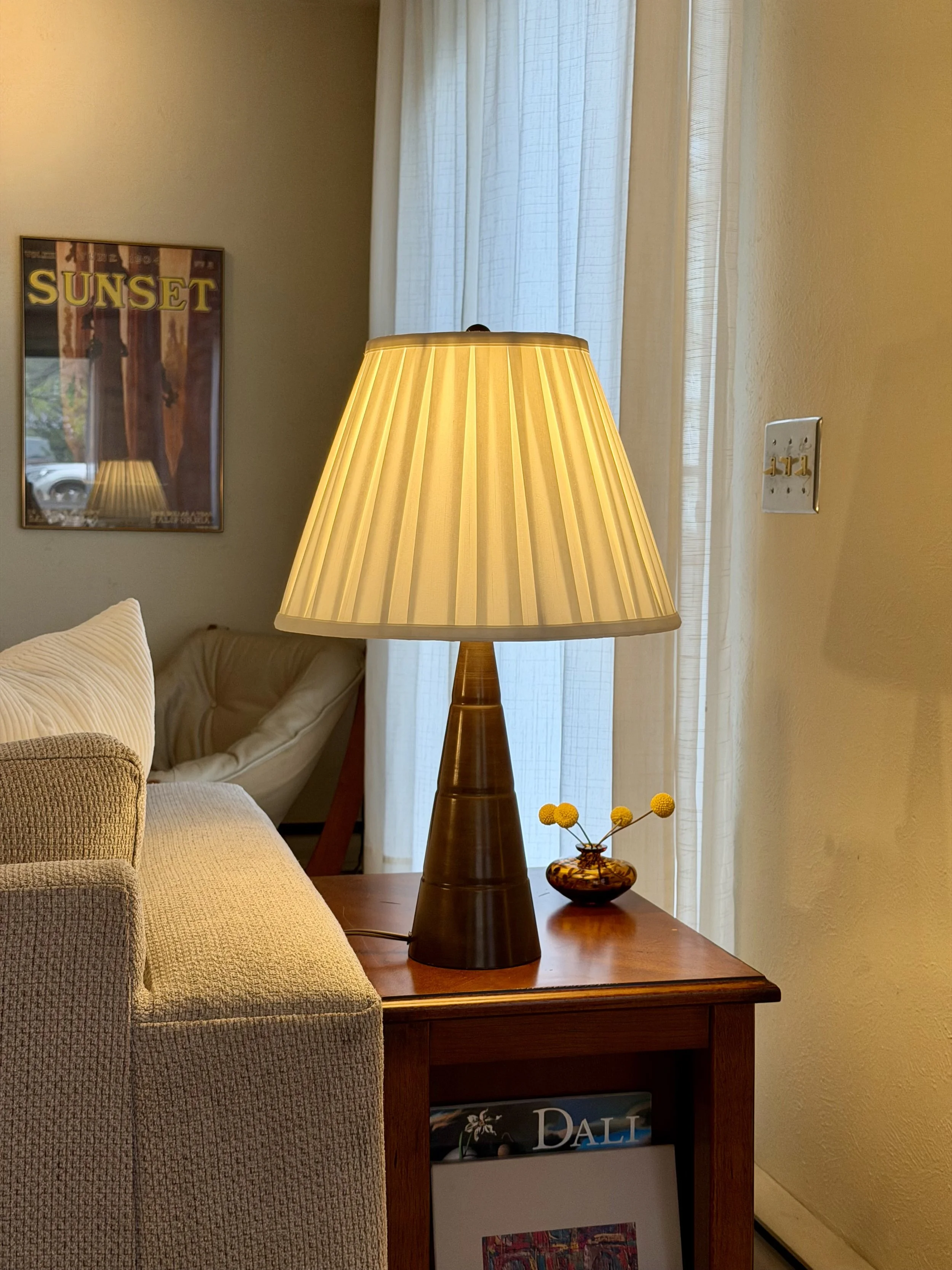 A living room scene with a beige sofa, a wooden side table, a vintage table lamp with a pleated shade, a small decorative vase with yellow flowers, a wall with a artwork labeled "SUNSET," and a window with light-colored curtains.