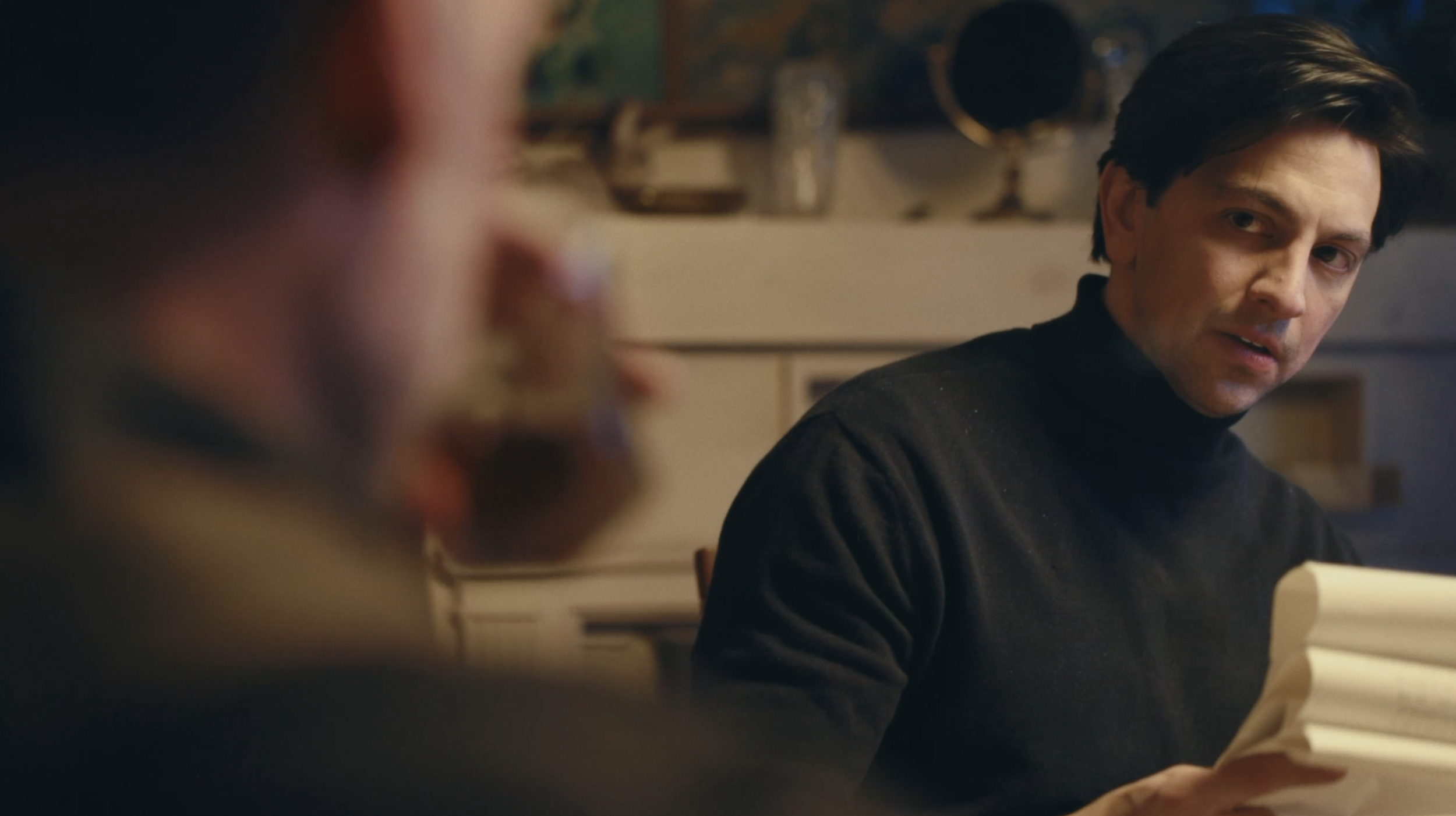 A man with dark hair wearing a black turtleneck, sitting at a table in a cozy room, speaking and looking at another person whose face is not visible.
