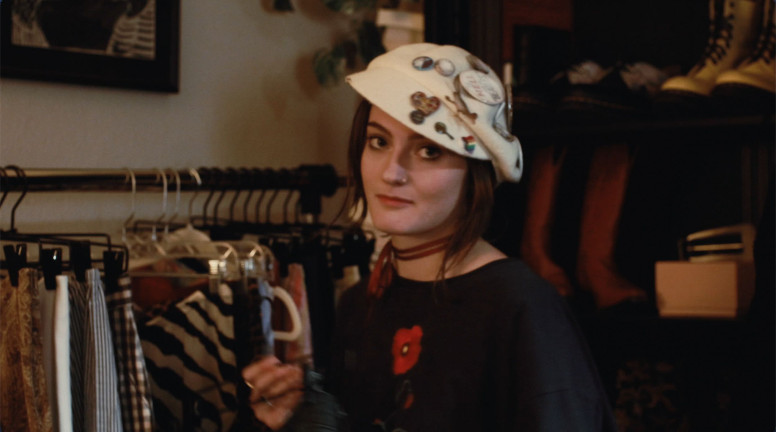 A woman with short brown hair looking at the camera, wearing a white hat decorated with various pins and buttons, and a black shirt with a red poppy. She is in a clothing store surrounded by racks of clothes.